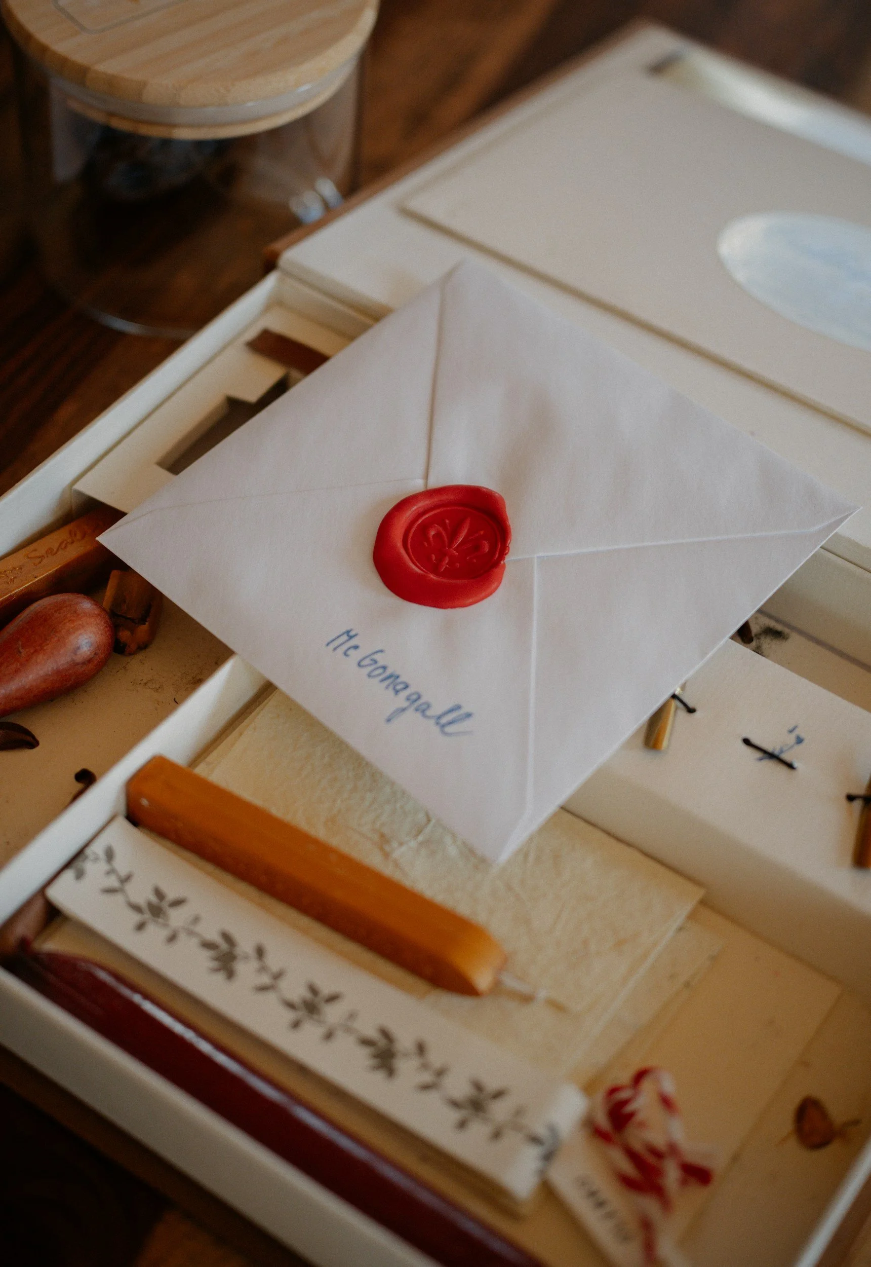 A white envelope with a red wax seal and handwritten message on the front, placed on a box with decorative items, including a heart-shaped object and ribbons.