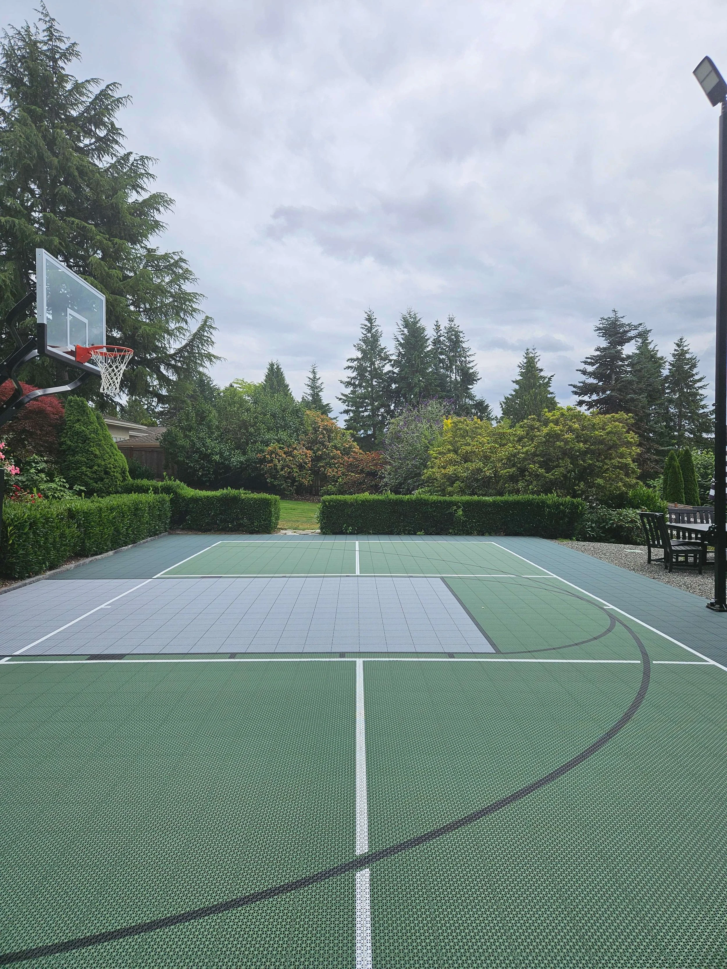 An outdoor basketball court with a green and gray surface, surrounded by trees and bushes, with a cloudy sky overhead.