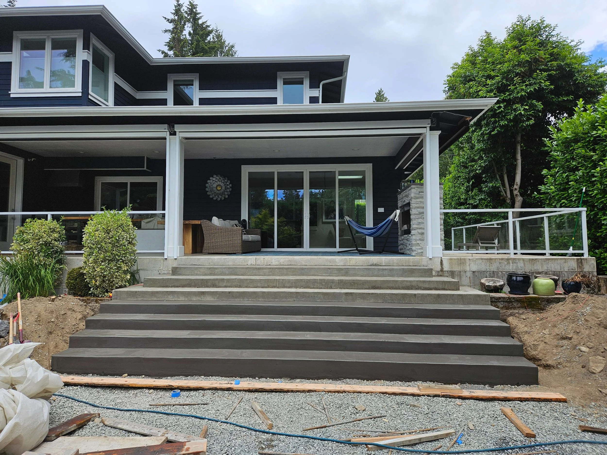 Backyard of a modern house under construction, with concrete stairs leading to a patio featuring outdoor furniture, a hammock, and a stone fireplace, surrounded by greenery and construction materials.