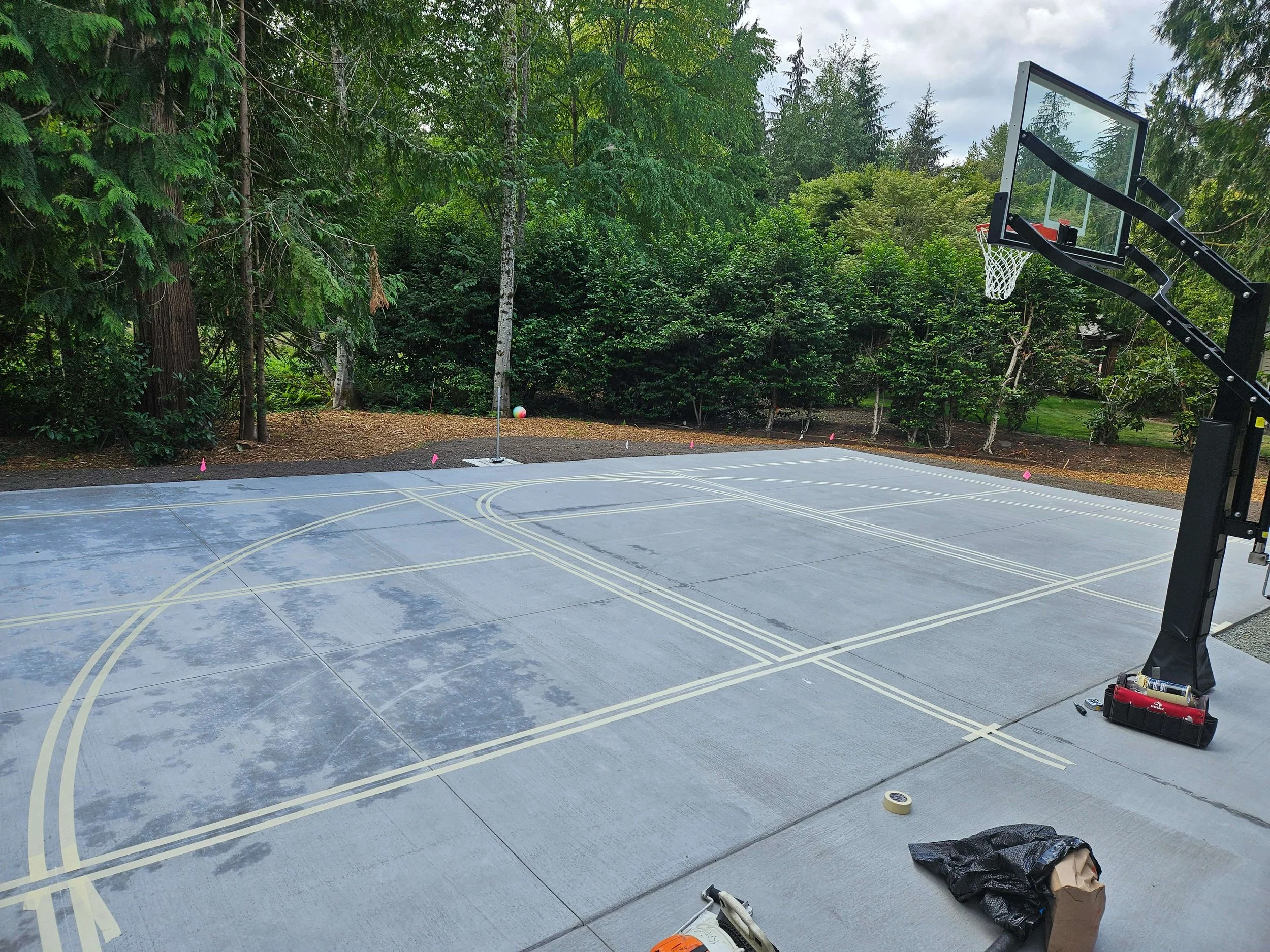 A newly painted outdoor basketball court with marked lines, secured with masking tape, surrounded by a forested area with tall trees.
