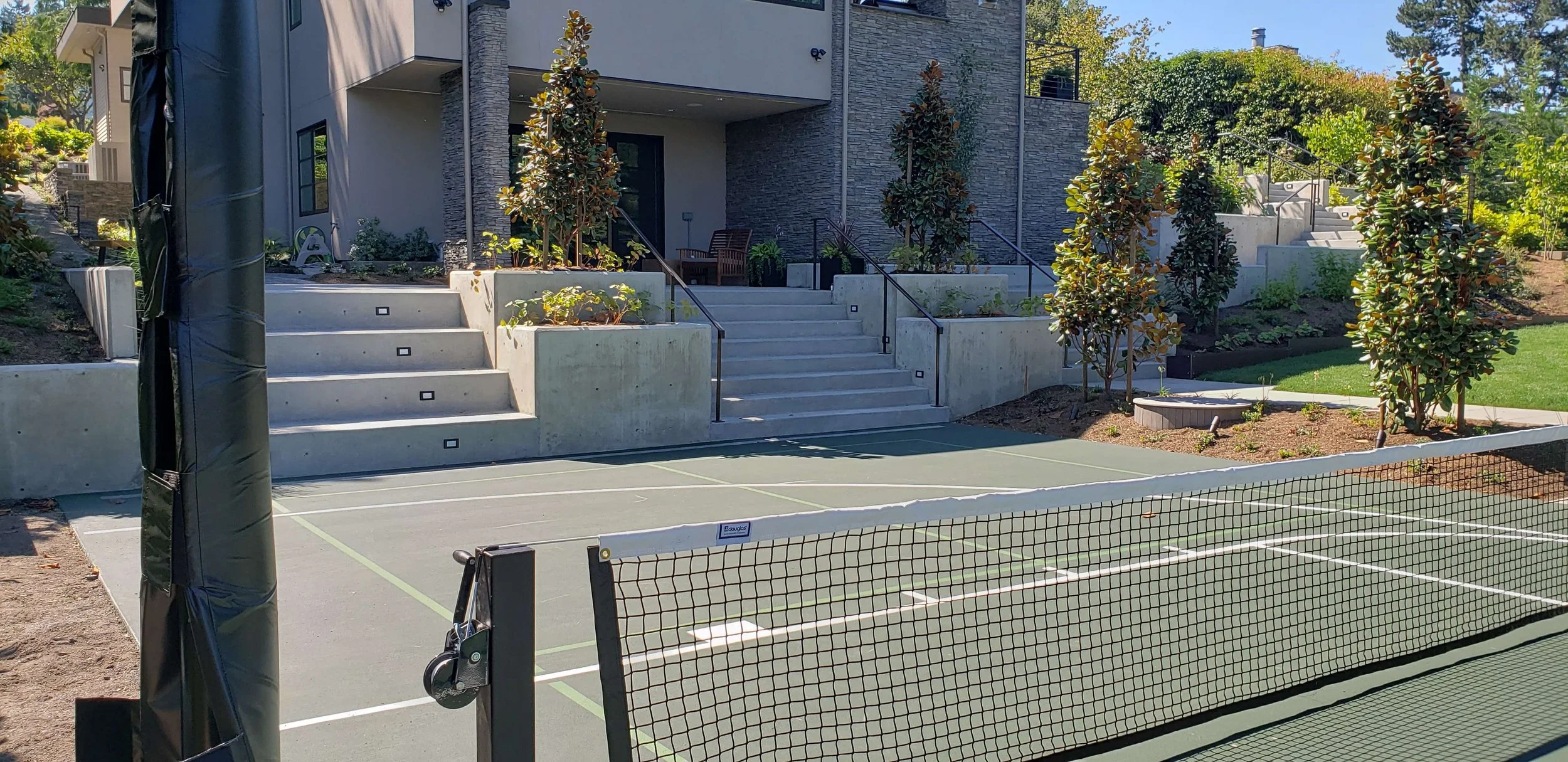 A modern house with a concrete staircase, landscaped with trees and plants, is adjacent to a tennis court with a net in the foreground, on a sunny day.