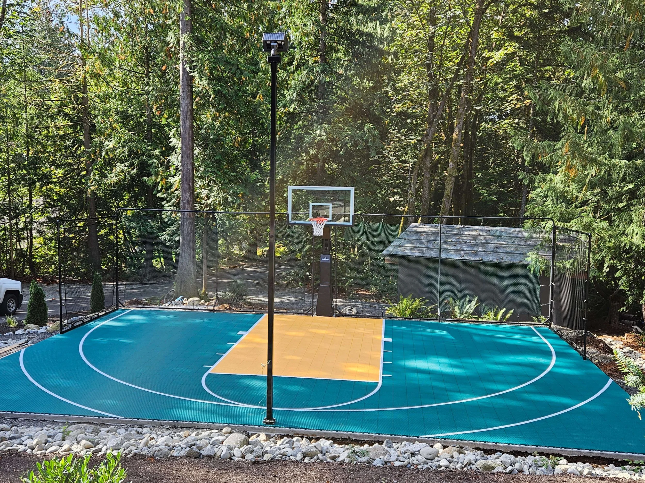Outdoor basketball court with a bright blue surface with white lines, a yellow painted area near the hoop, a black backboard with a white rim, surrounded by a black safety fence, trees in the background, and a small shed on the right side.