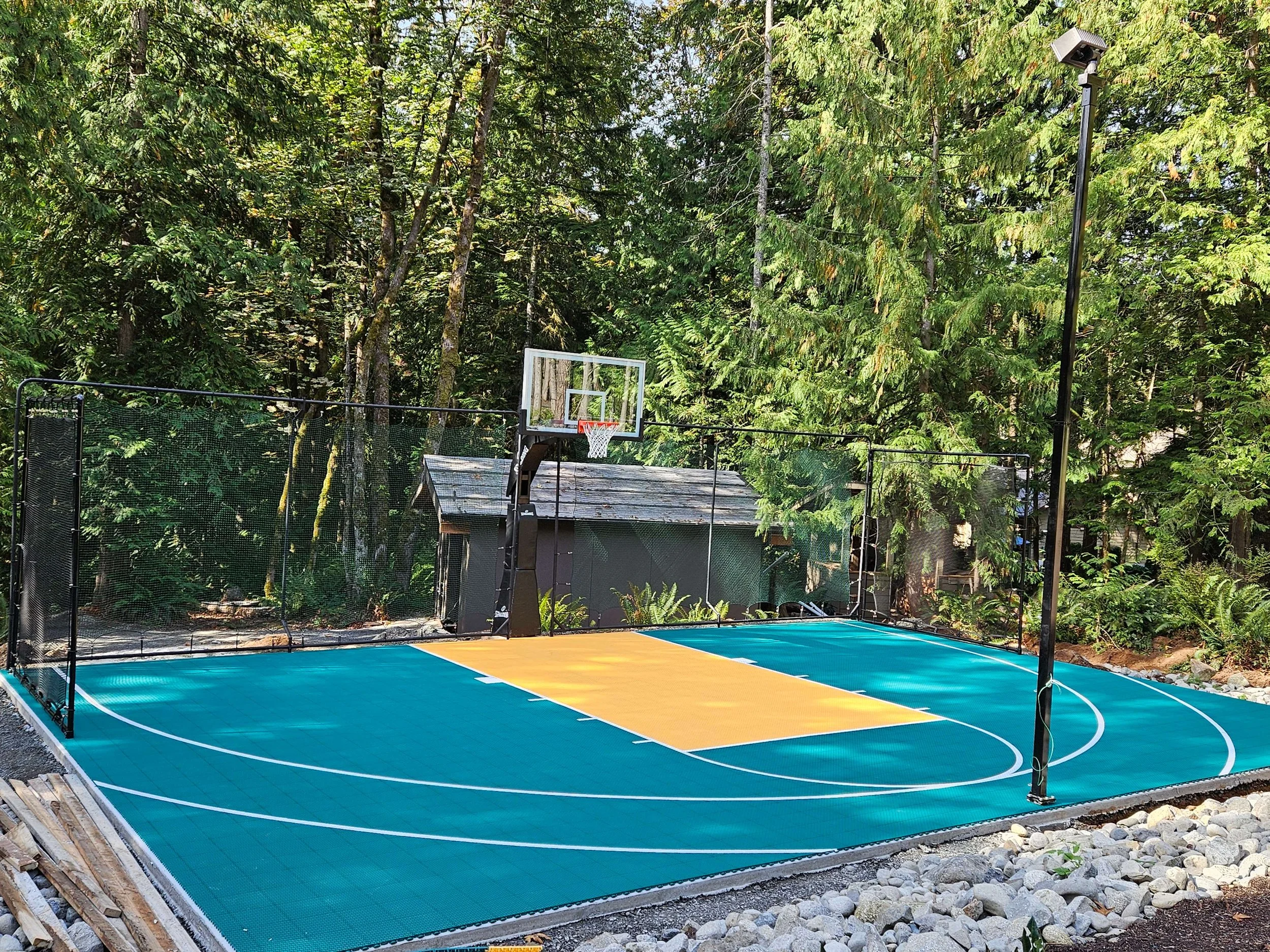 Outdoor miniature basketball court with a green, yellow, and blue surface, a basketball hoop, and a netted fence, surrounded by tall trees.