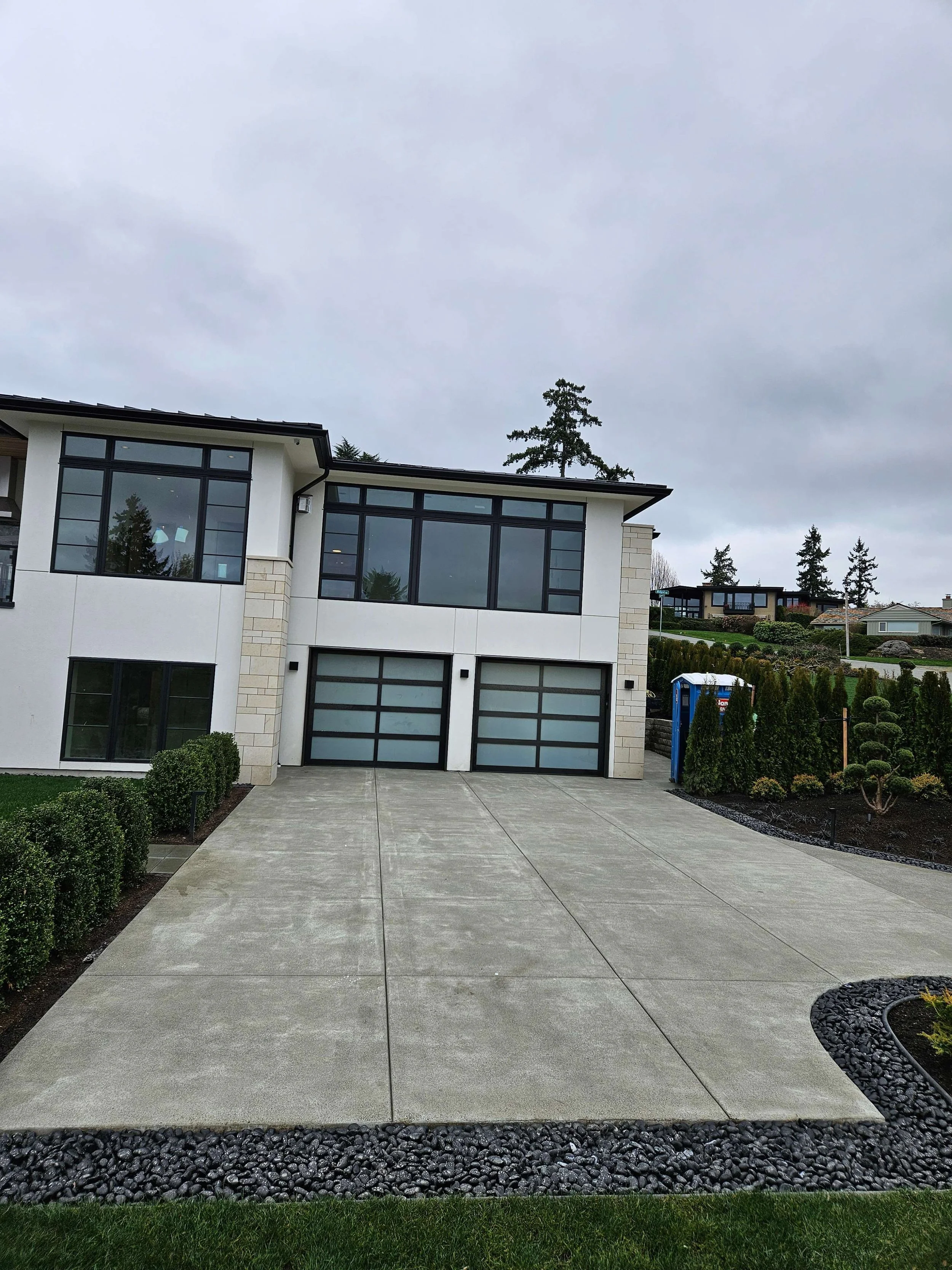 Modern two-story house with large windows, a concrete driveway, and surrounding shrubbery under an overcast sky.