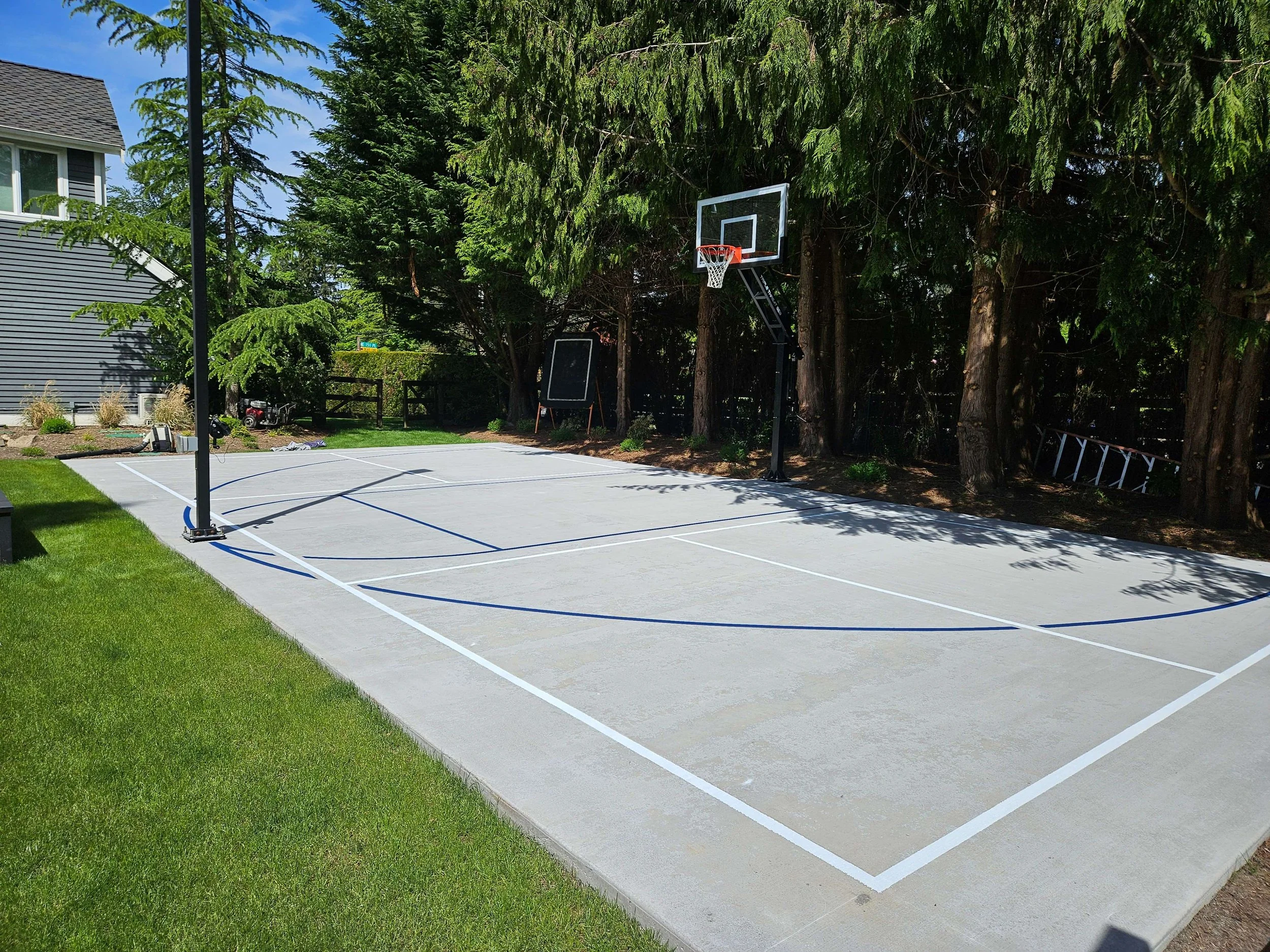 A newly painted outdoor basketball court with white lines and two basketball hoops, surrounded by green grass and tall trees.