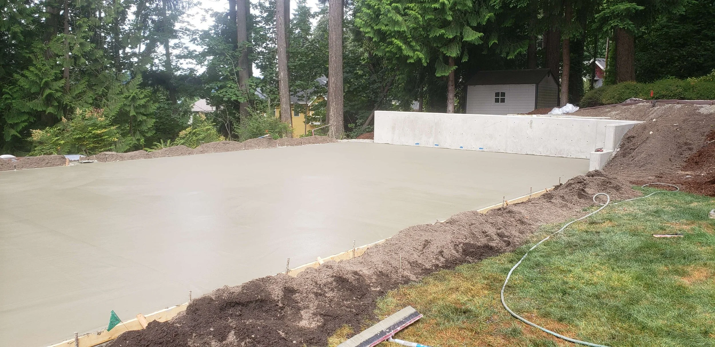 Freshly poured concrete slab foundation for a construction project in a backyard, surrounded by dirt and construction tools.