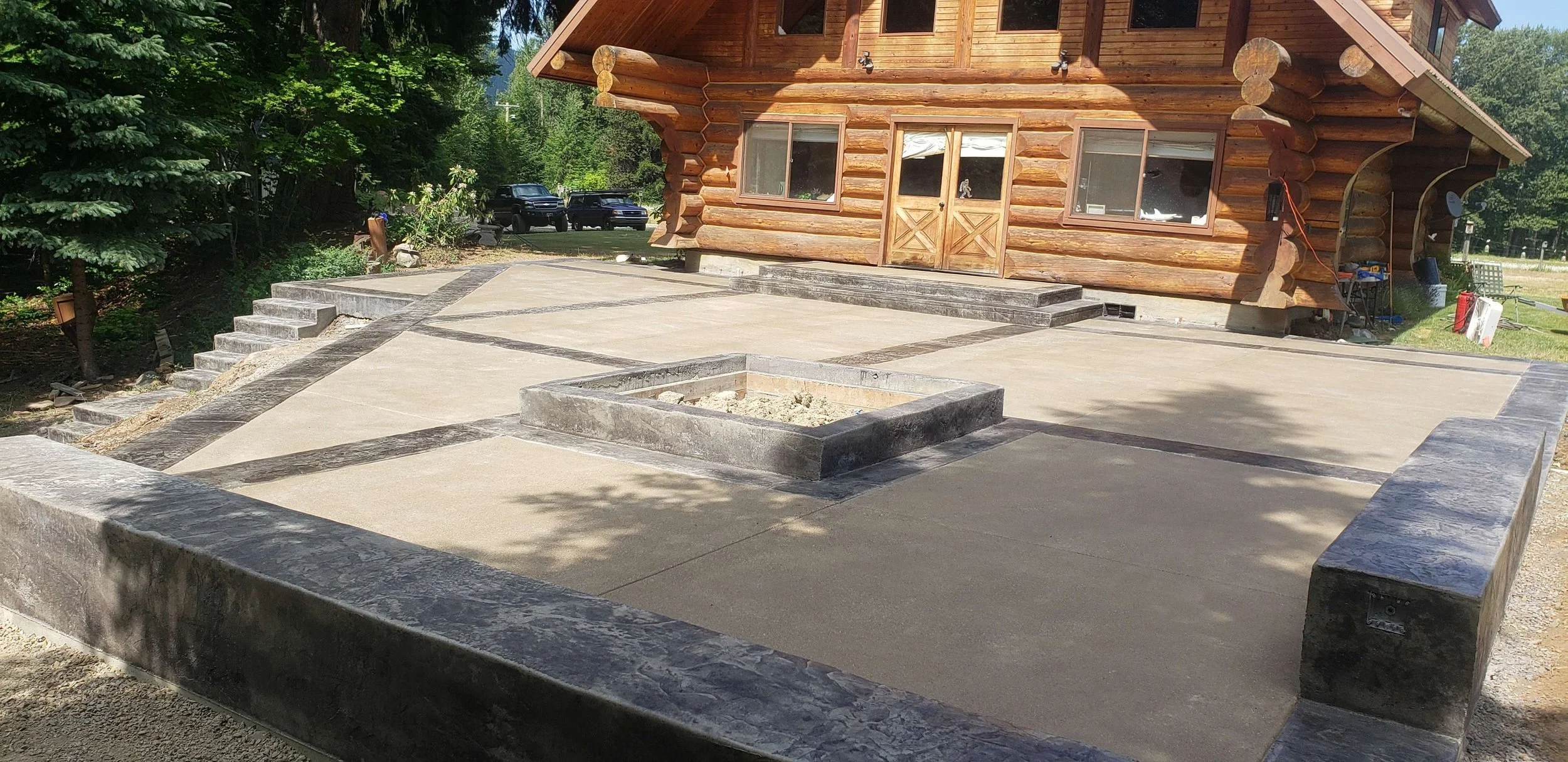 Constructed concrete patio with stone borders in front of a wooden cabin, with a small raised square feature in the center.
