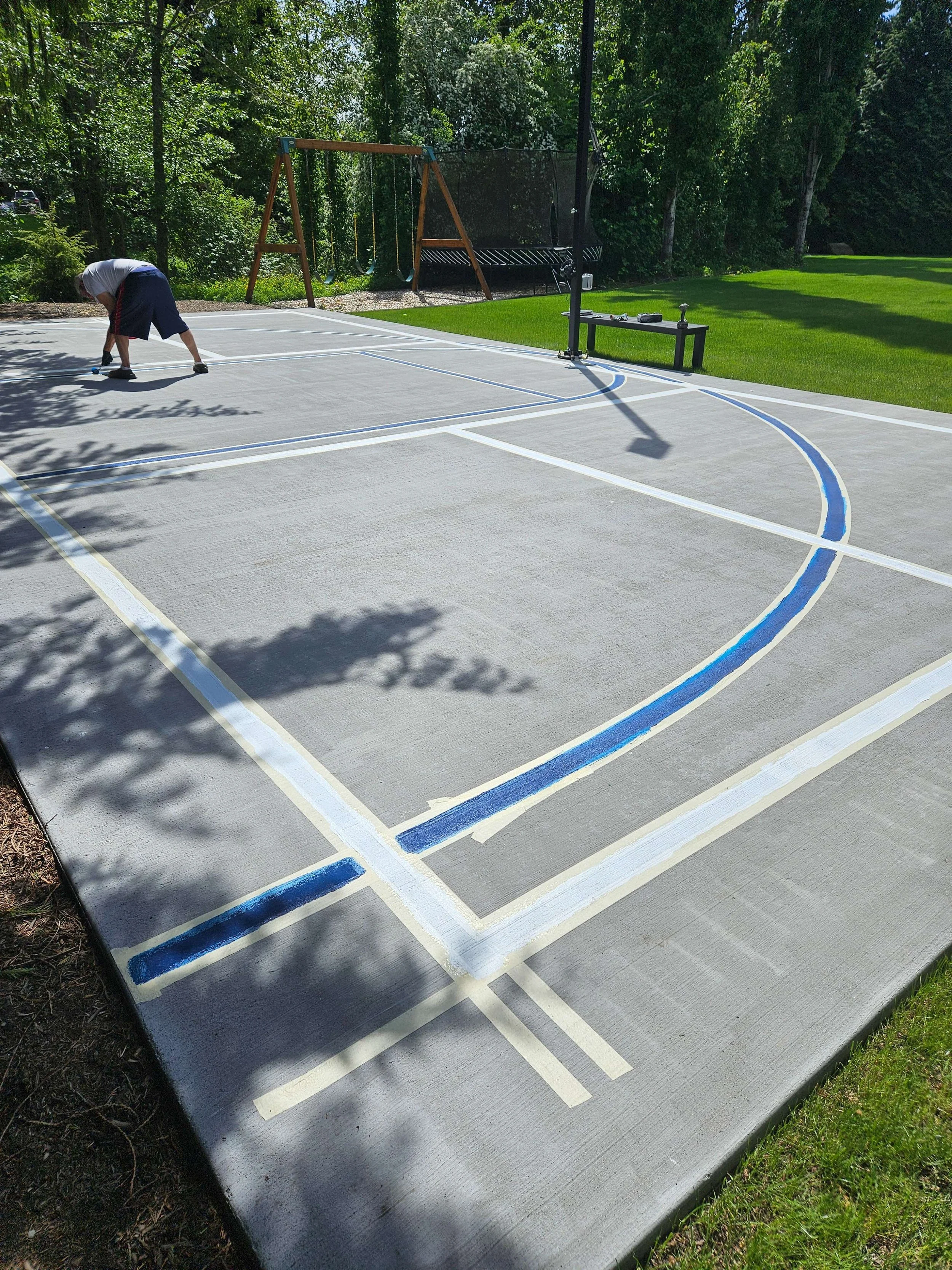 A person working on painting lines on a basketball court in a backyard, with trees and greenery surrounding the area and a swing set in the background.
