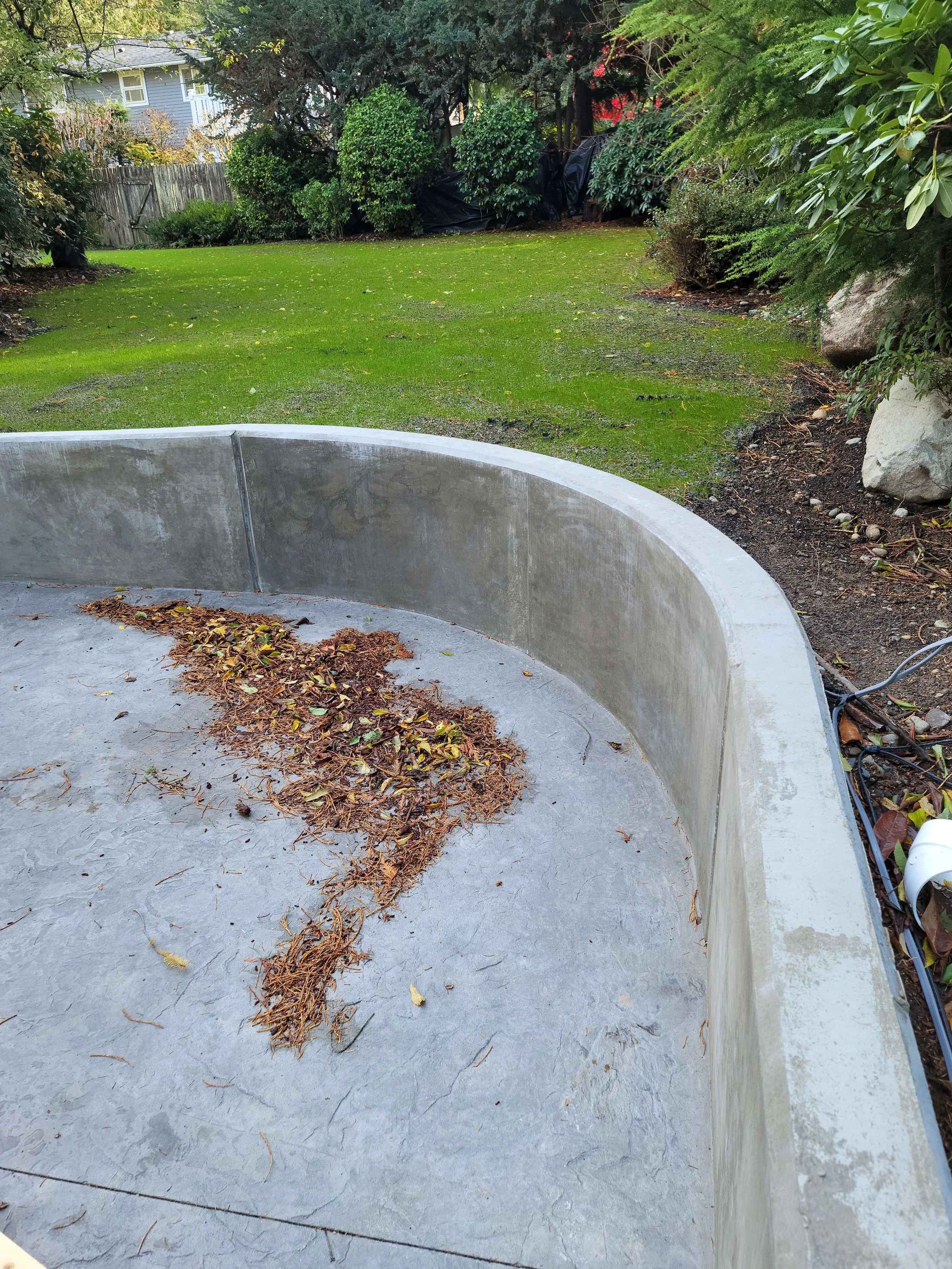 A backyard with a well-maintained lawn, trees, and shrubs. In the foreground, part of a round concrete structure with dried leaves and debris on the surface.