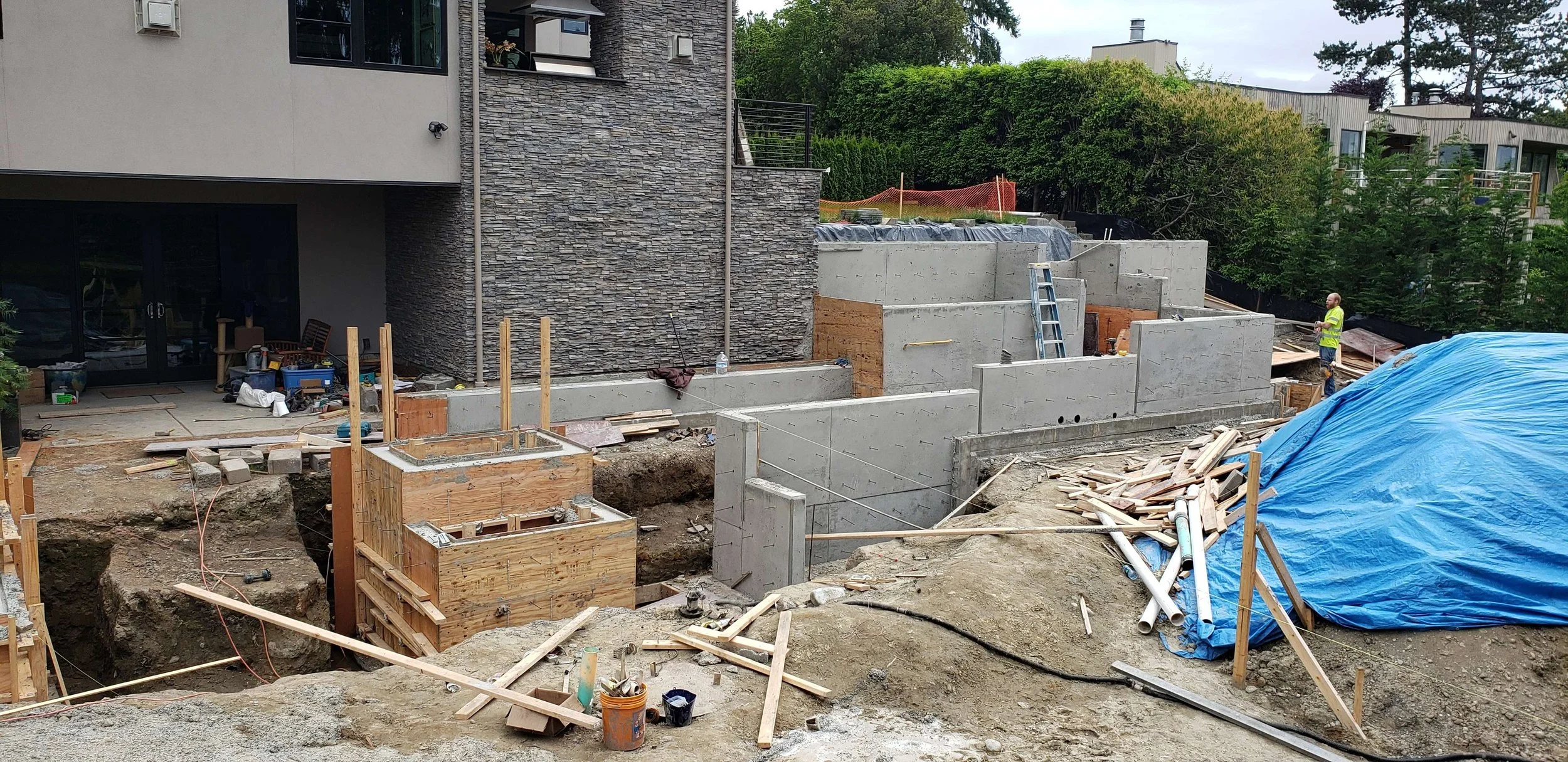 Construction site with workers, concrete walls, wooden forms, pipes, and a blue tarp covering materials.