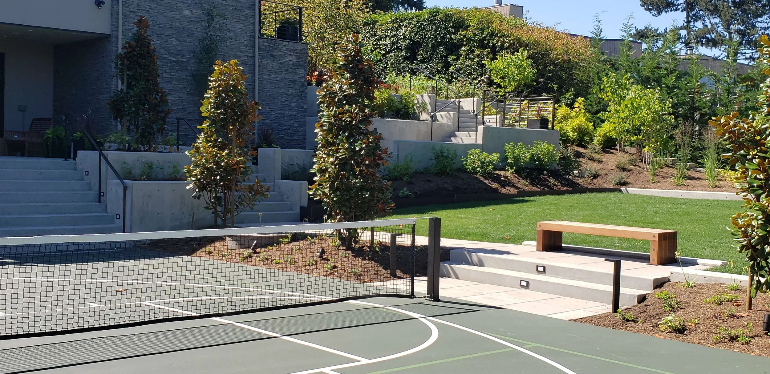 A modern outdoor scene featuring a tennis court with a net, a wooden bench, wide stairs, and landscaped greenery with trees and bushes in the background.