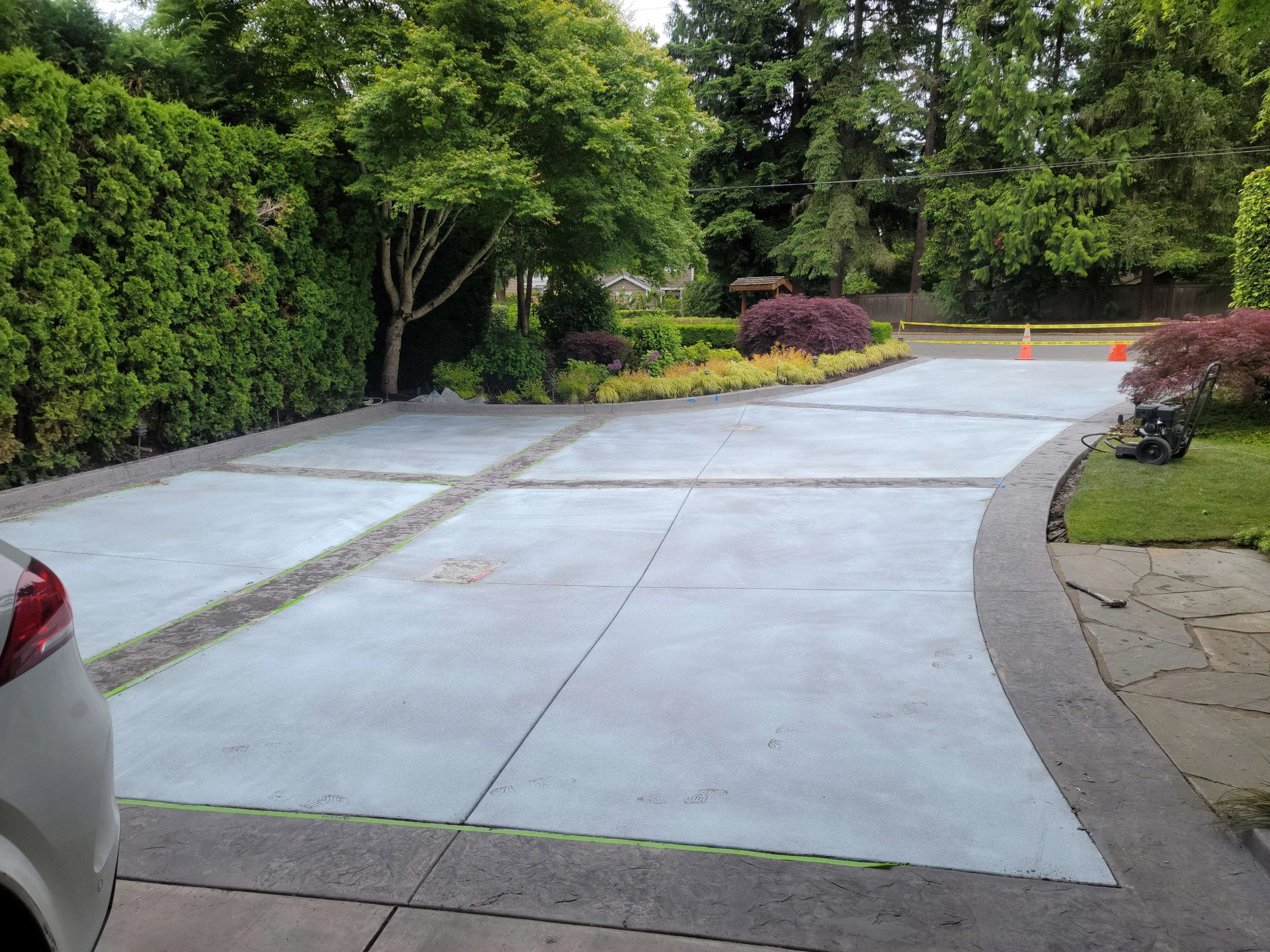 A driveway covered with a light grey liquid, possibly concrete or paint, with visible handprints and footprints. The driveway is bordered by greenery, bushes, and trees, and there are orange cones and caution tape at the end of the driveway indicatin
