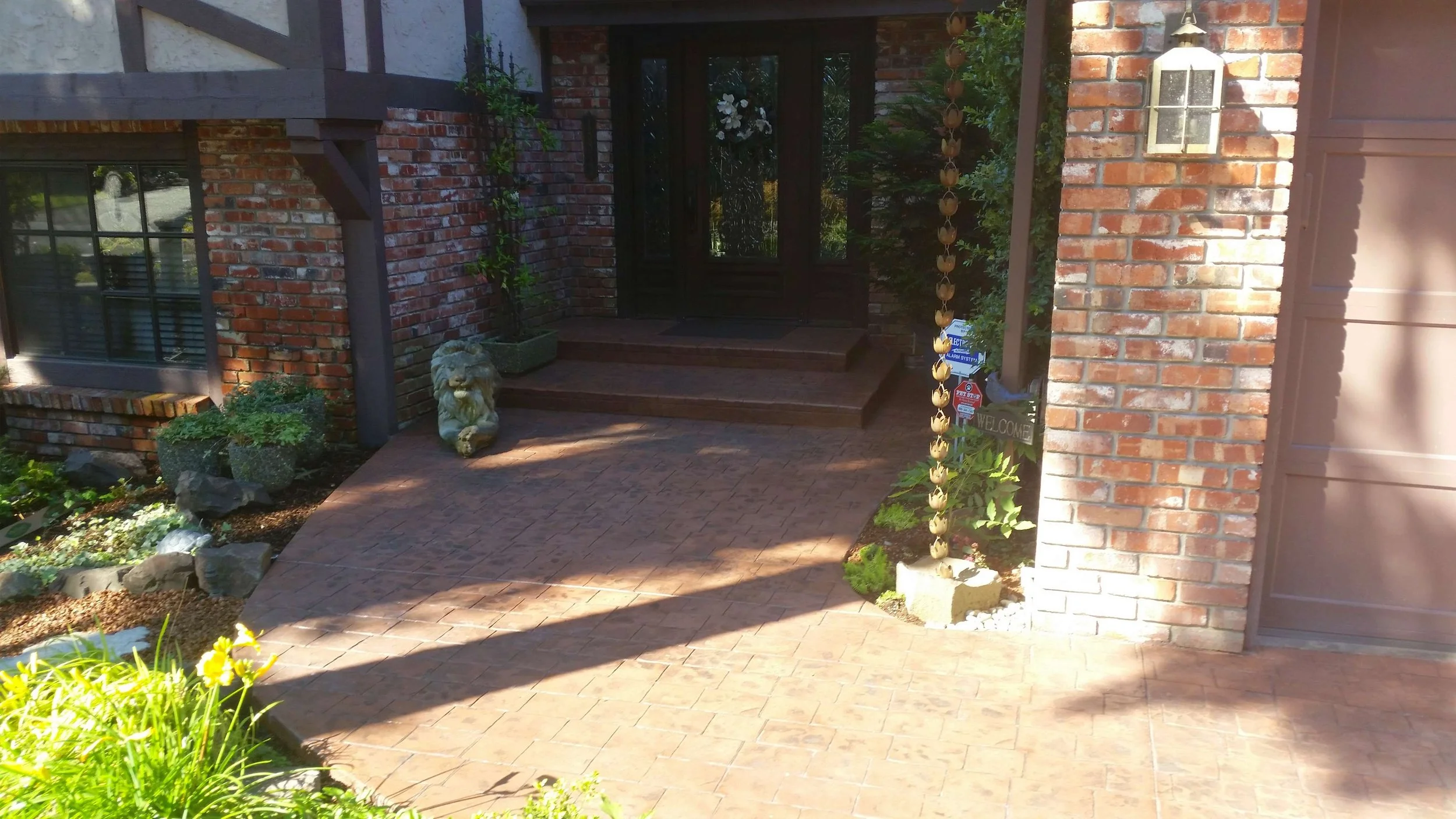 Front porch with brick steps, lion statue, potted plants, decorative metal wind chime, and welcome sign.