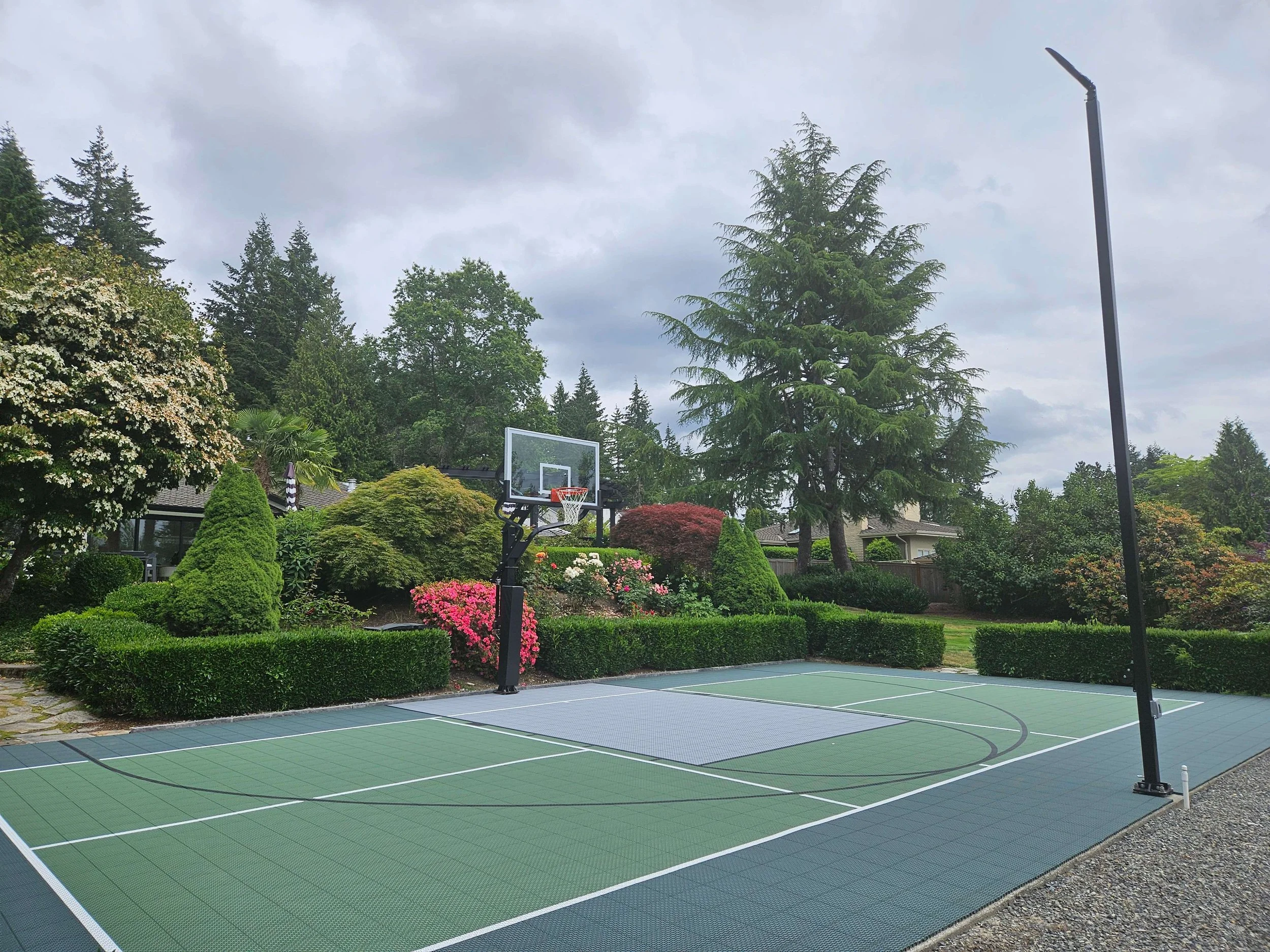 An outdoor basketball court surrounded by lush green trees and shrubs, with a net and hoop, in a residential area under a cloudy sky.