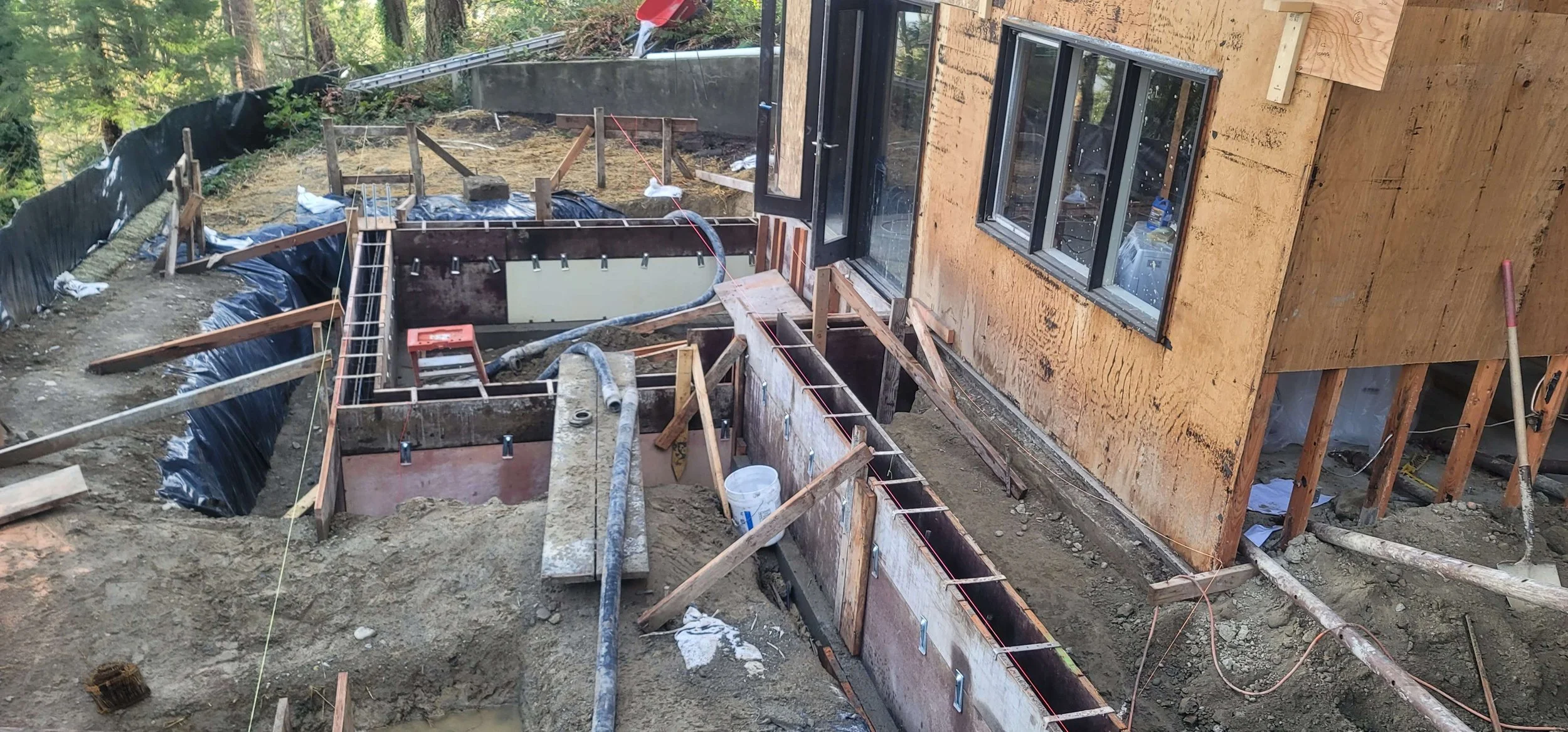 Construction site of a house extension with foundation work and framing in progress, showing exposed wooden beams, concrete forms, scaffolding, and building materials, adjacent to an existing building with windows.