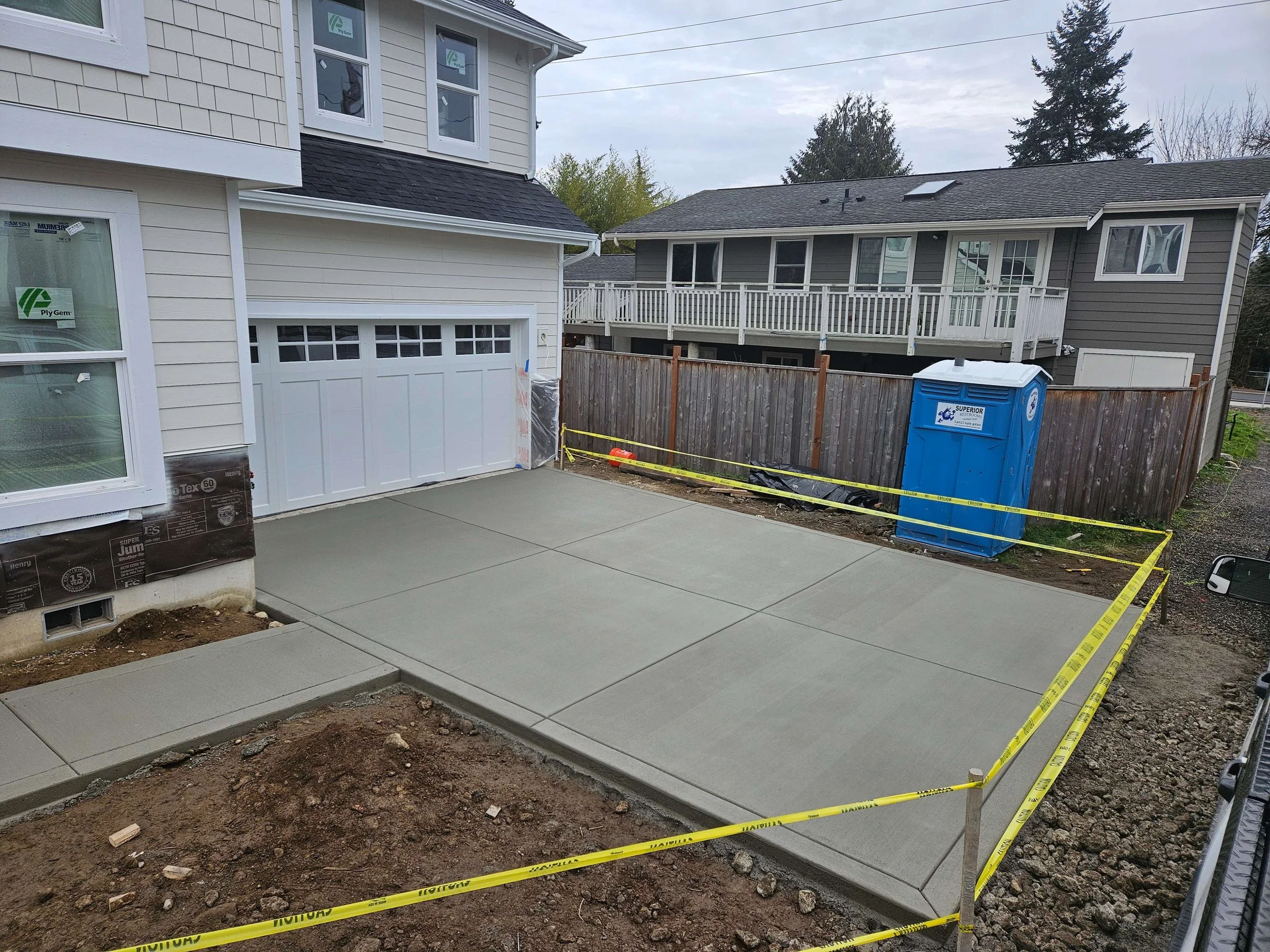 Newly poured concrete driveway in front of a house, surrounded by yellow caution tape, with a blue portable toilet in the yard.