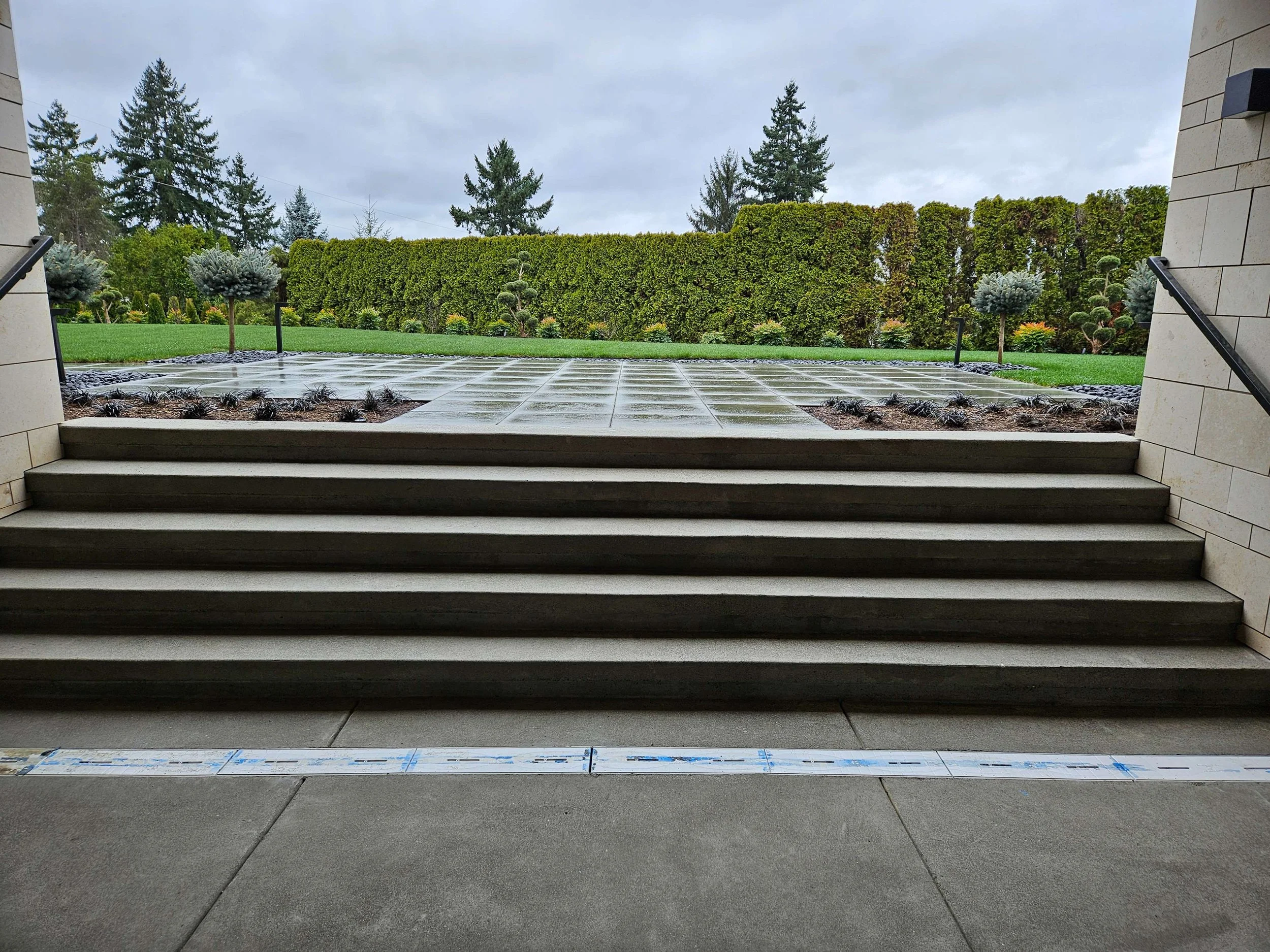 View from a building staircase overlooking a landscaped garden with decorative bushes, trimmed hedge, and a wet tiled pathway on a cloudy day.
