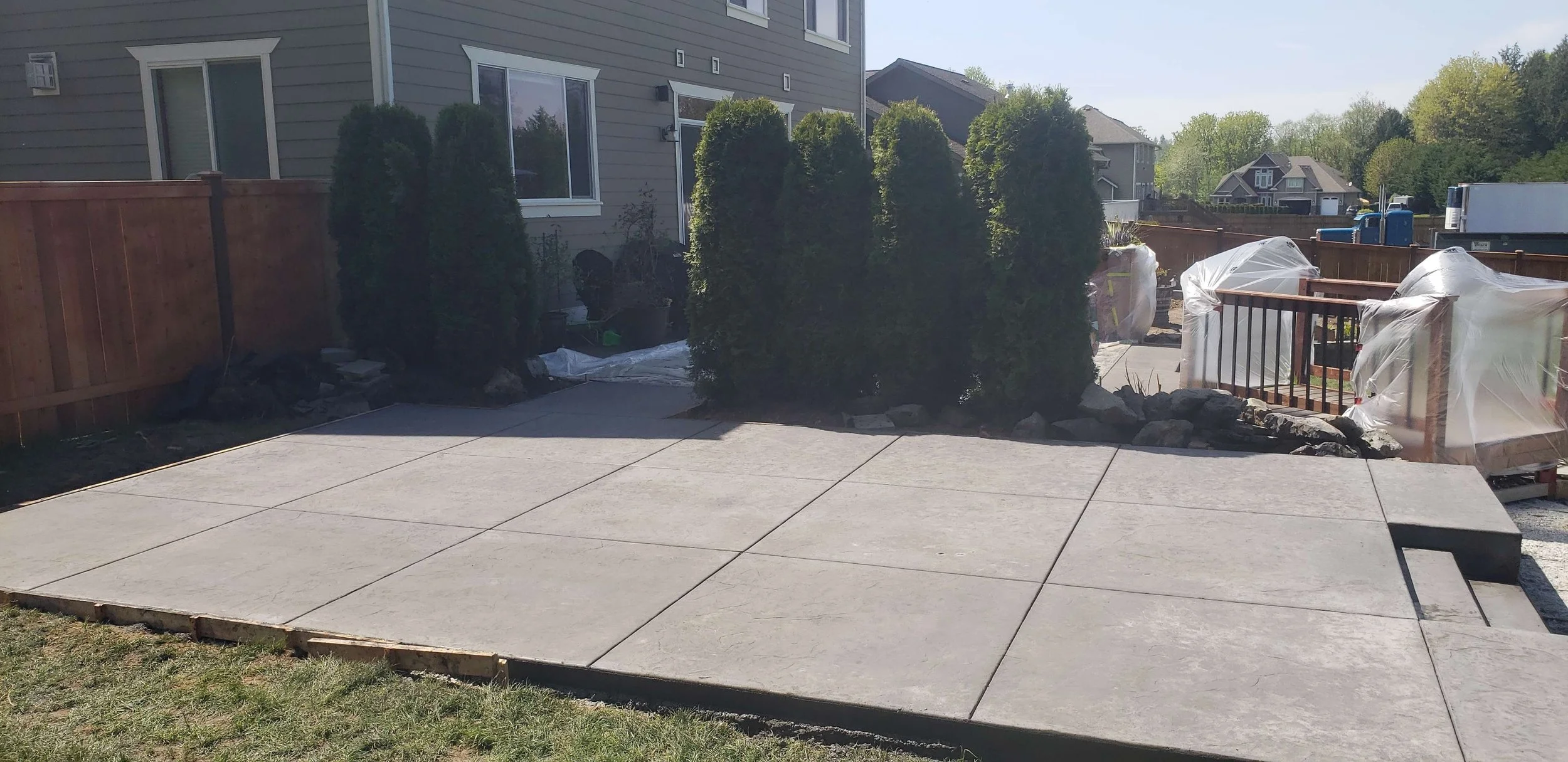 A newly poured concrete patio in a backyard next to a house with large windows. The patio is bordered by a wooden fence on one side and has some bushes and rocks around the edges. There are construction materials covered in plastic on the deck, with
