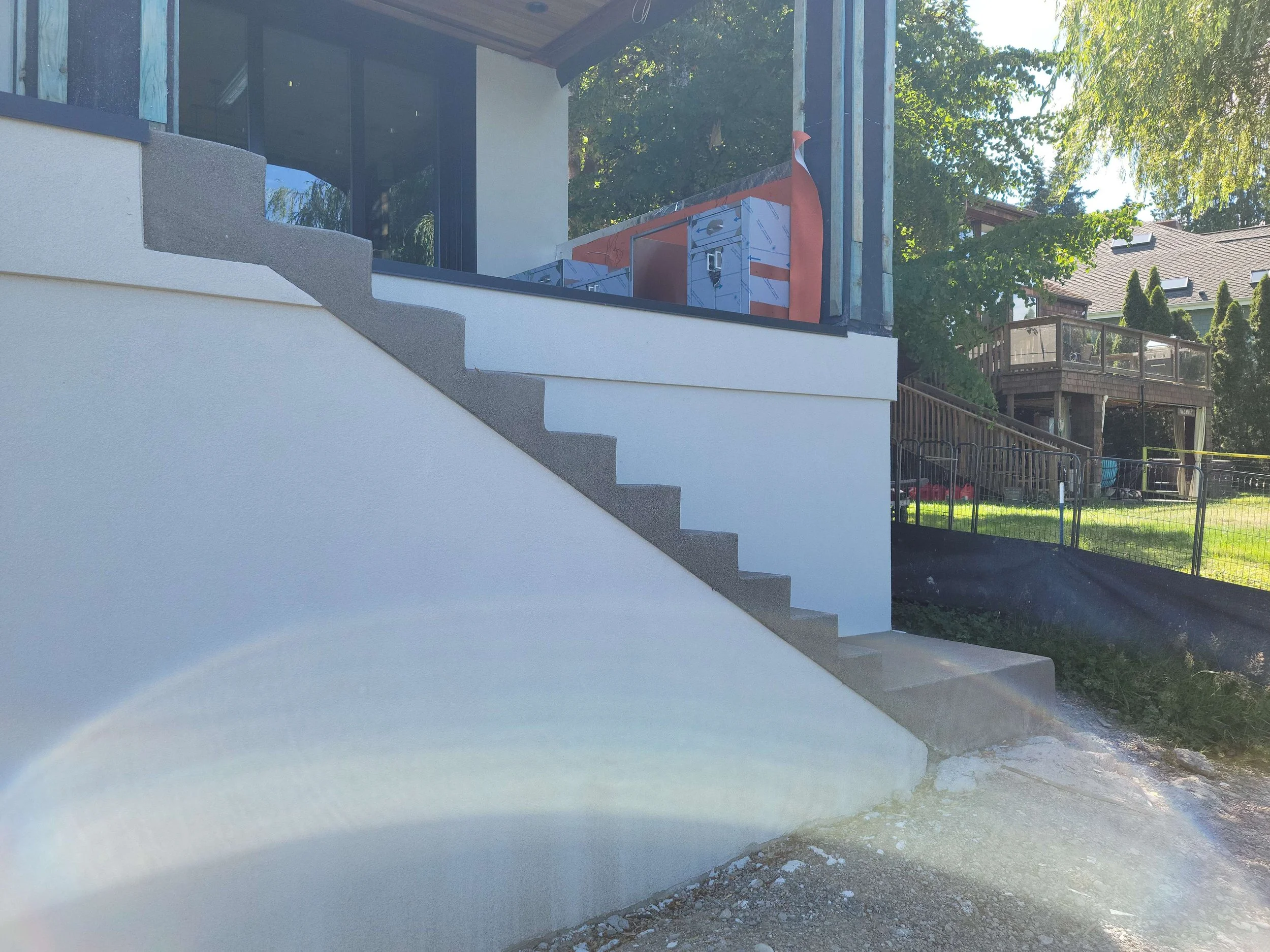 View of a modern house with an outdoor staircase, glass walls, and construction materials visible on a balcony, surrounded by trees and neighboring houses.