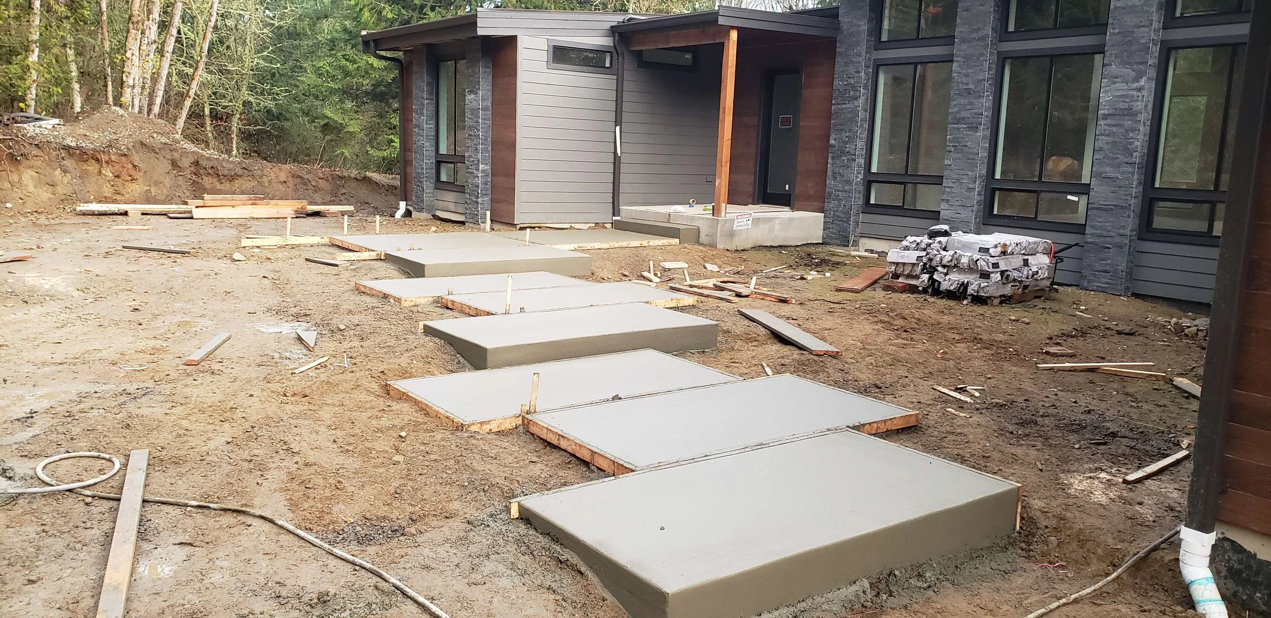 Construction site with newly poured concrete slabs forming the walkway in front of a modern house with gray siding, large windows, and a wood accent porch. Piles of lumber and construction materials are scattered around.