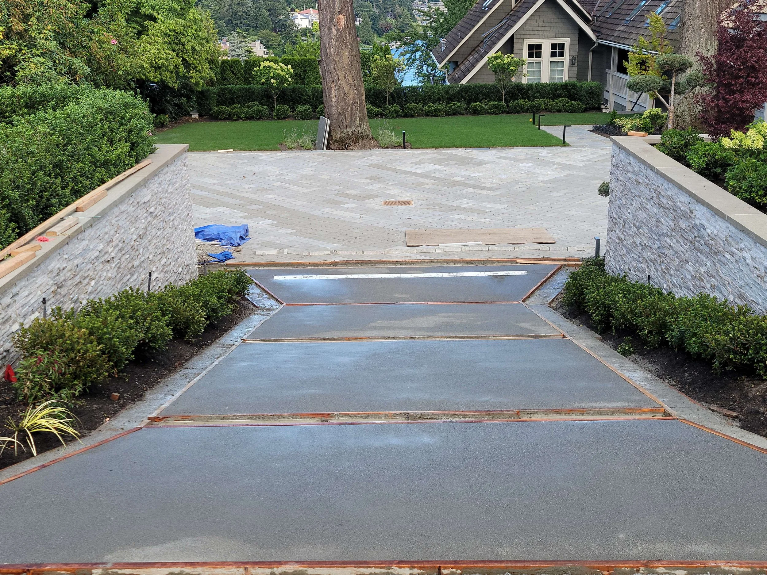 A driveway under construction with rebar and concrete sections, leading from a landscaped yard with bushes to a paved area with stones, houses, and trees in the background.