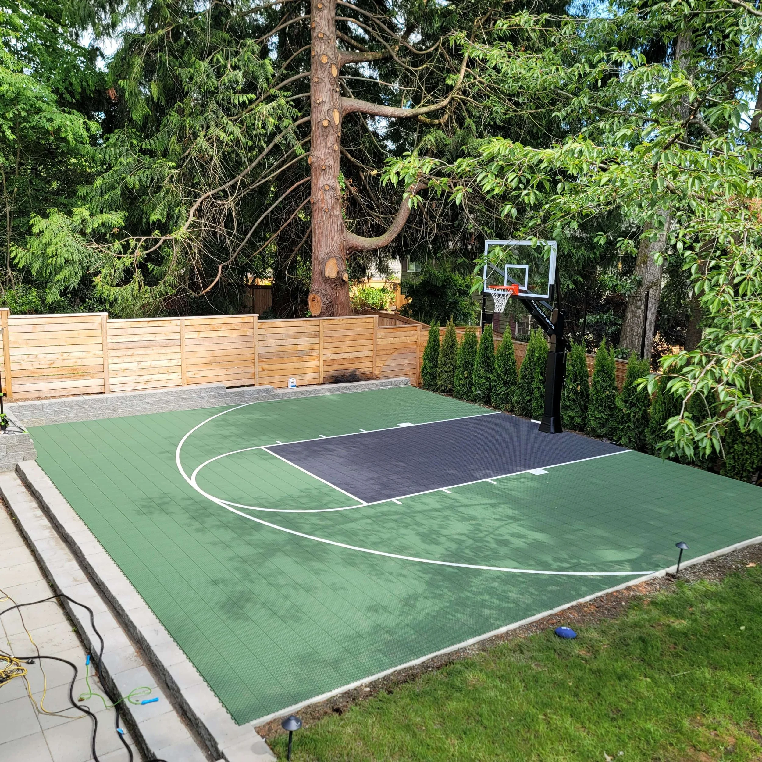 A newly installed outdoor basketball court with a black and green surface, surrounded by a wooden fence and lush green trees.