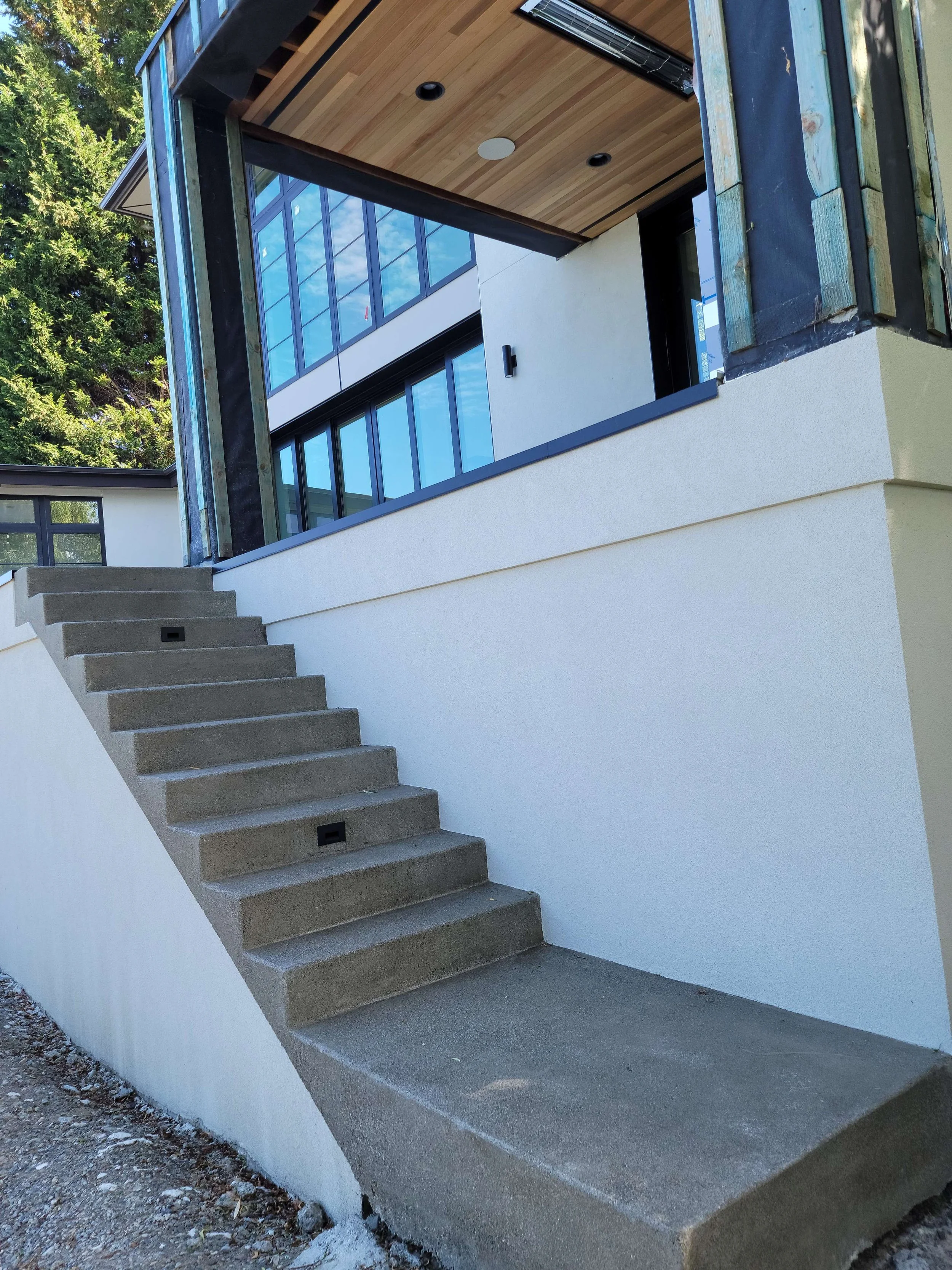 Exterior of a modern house with concrete stairs leading up to a balcony with large glass windows, wooden ceiling, and partial construction materials on the right side.