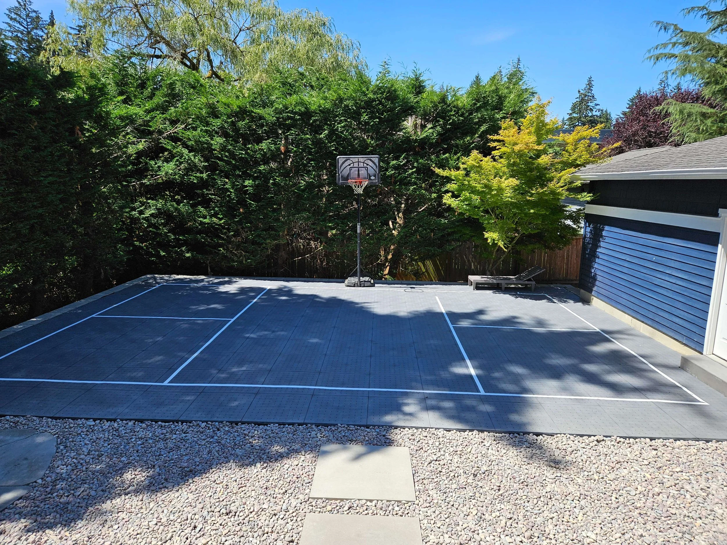 Outdoor backyard basketball court with a basketball hoop, surrounded by trees and a blue building on the right, with a lounge chair nearby.