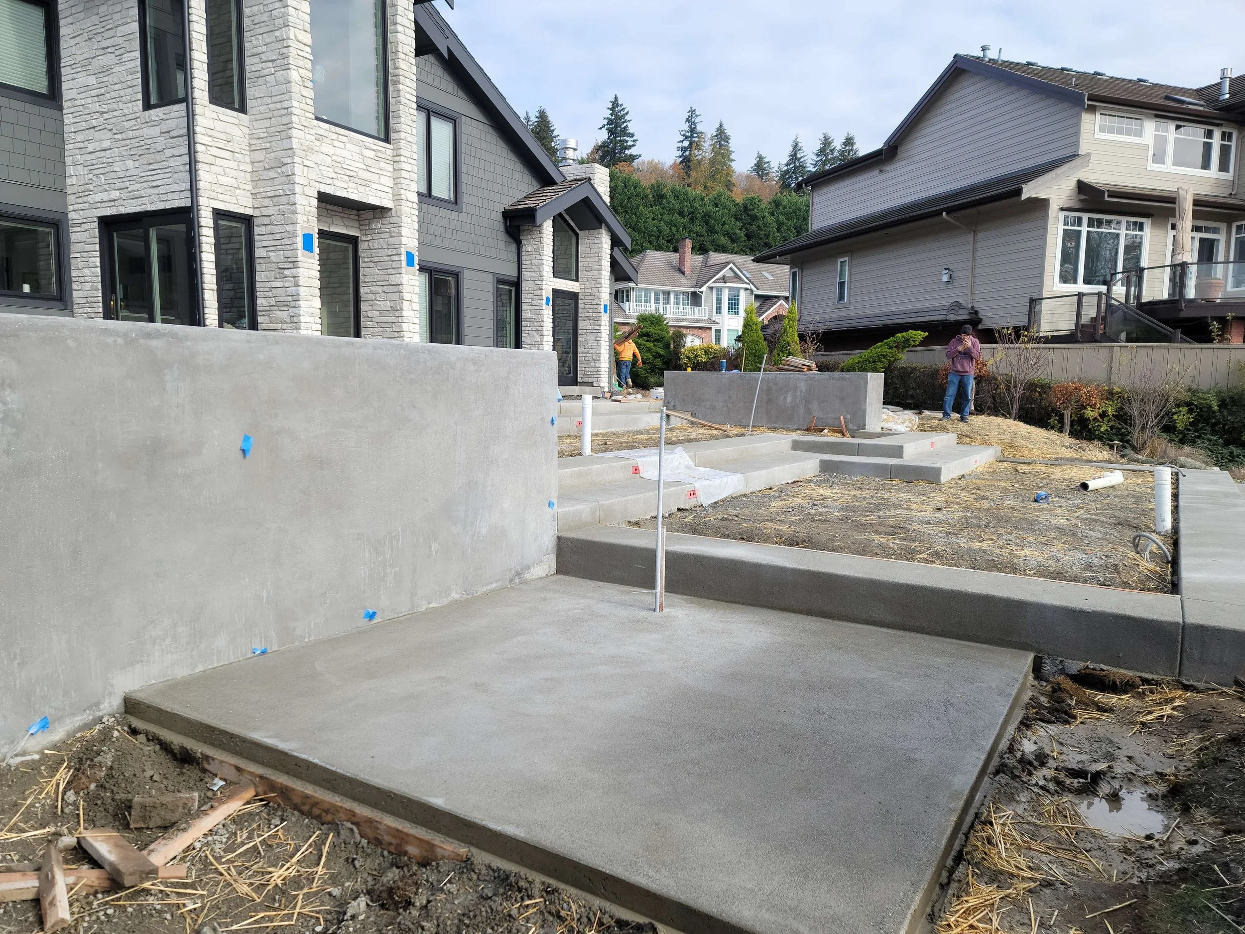 Construction site of a residential driveway with freshly poured concrete, concrete walls, and steps, surrounded by residential houses and workers.