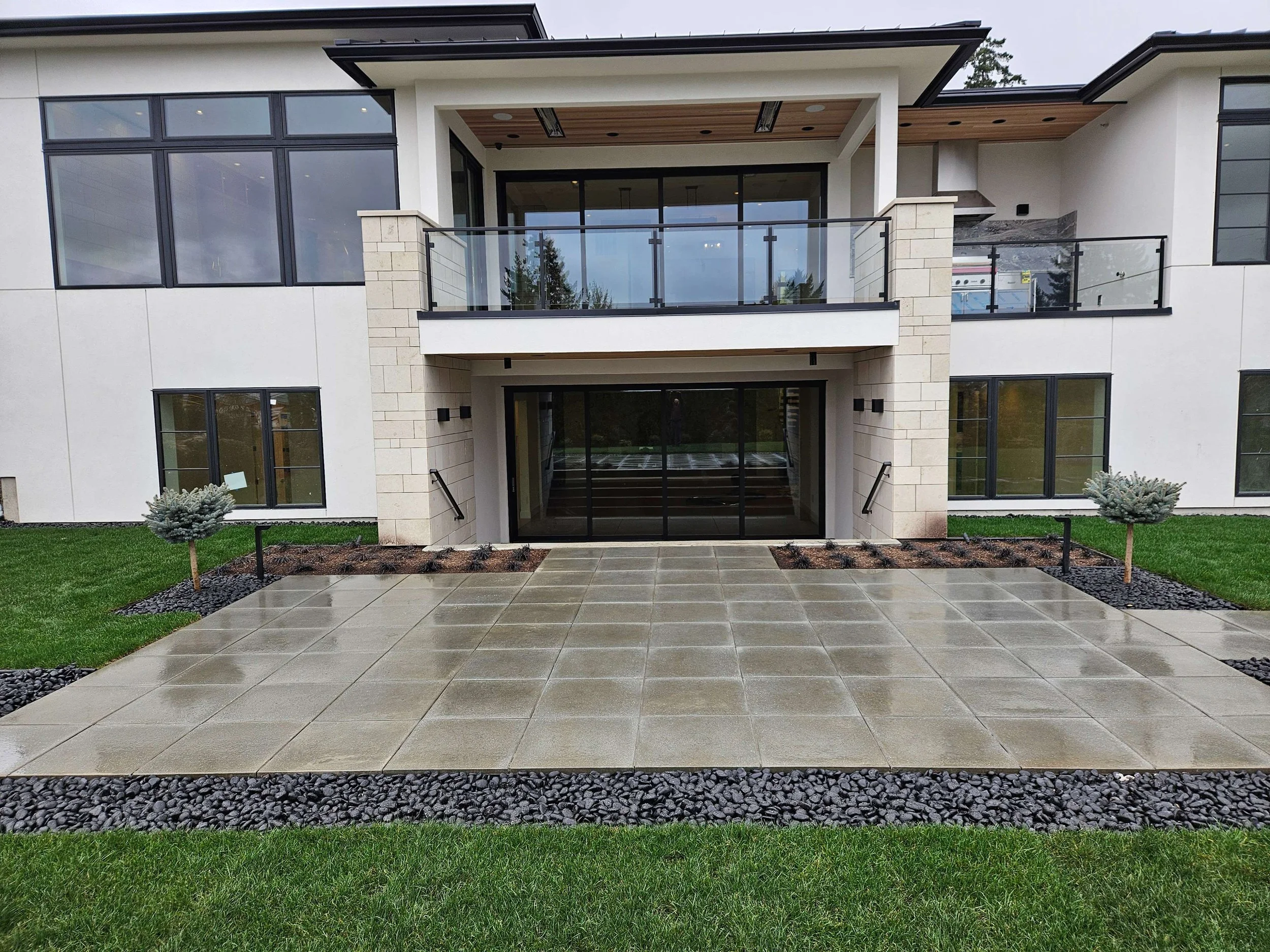 Front view of a modern two-story apartment building with a glass sliding door entrance, balcony, large windows, small landscaped garden with small trees, and a tiled walkway.