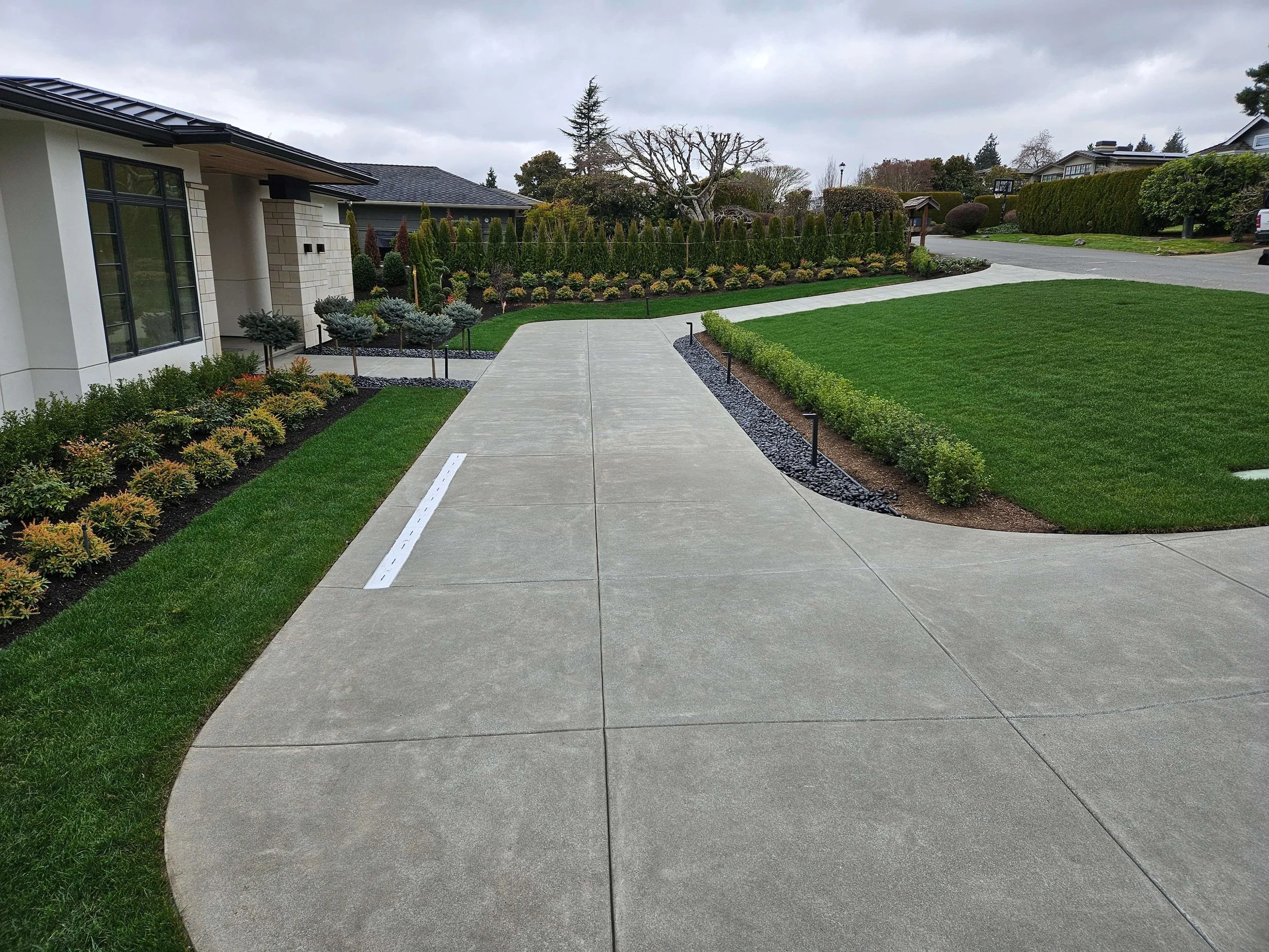 Clean concrete driveway and sidewalk in front of modern home with manicured lawn, plants, and bushes, on a cloudy day.