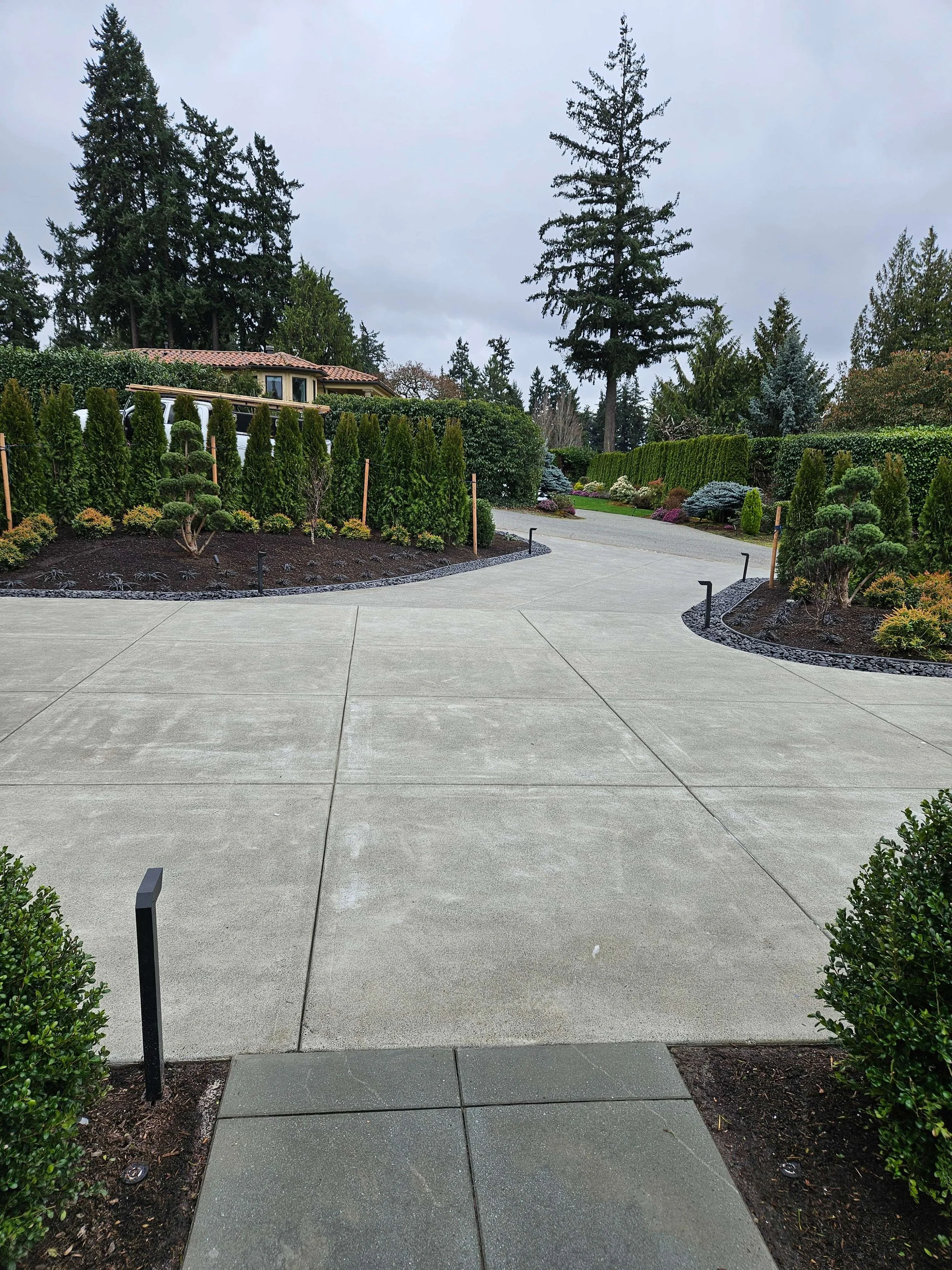 A paved driveway winding through a landscaped garden with tall green hedges and a variety of trees, overcast sky.