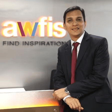 A man in a black suit and red tie standing in front of a wall with a logo that says 'awfis' and the words 'FIND INSPIRATION' below it, in an office setting.