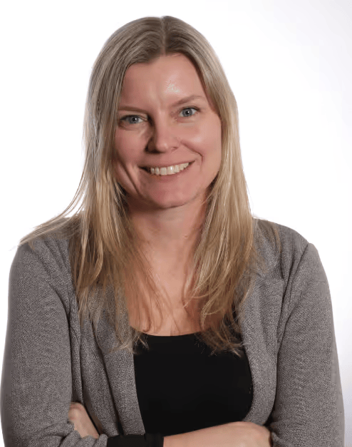 A woman with long blonde hair smiling, wearing a gray cardigan over a black top, standing against a white background.