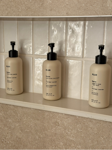 Three bottles of KLUR brand shampoo and hand soap on a bathroom shelf in front of a mirror and white tiled wall.