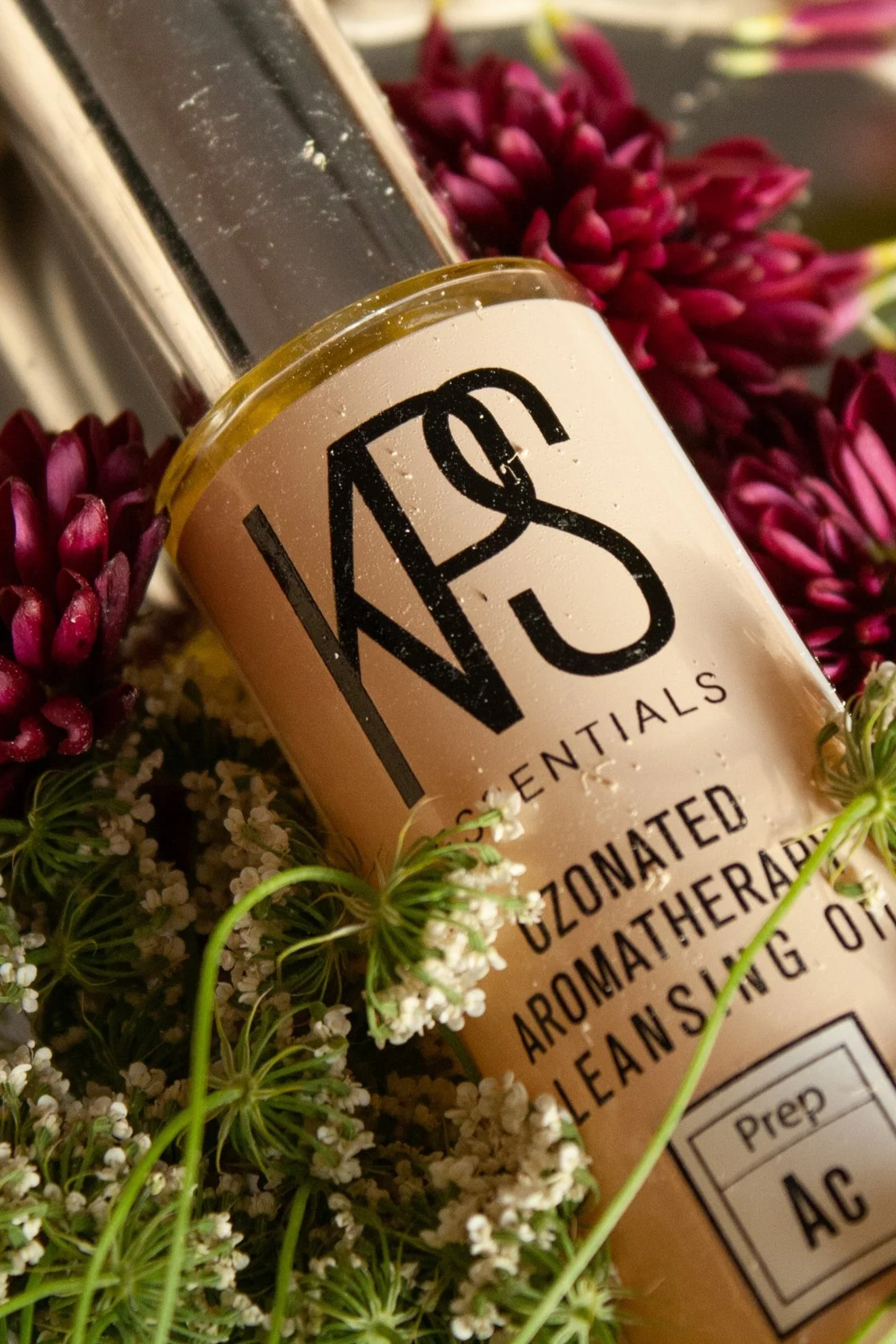 A bottle of essential oil labeled 'KPS' surrounded by pink, purple, and white flowers and green plants.