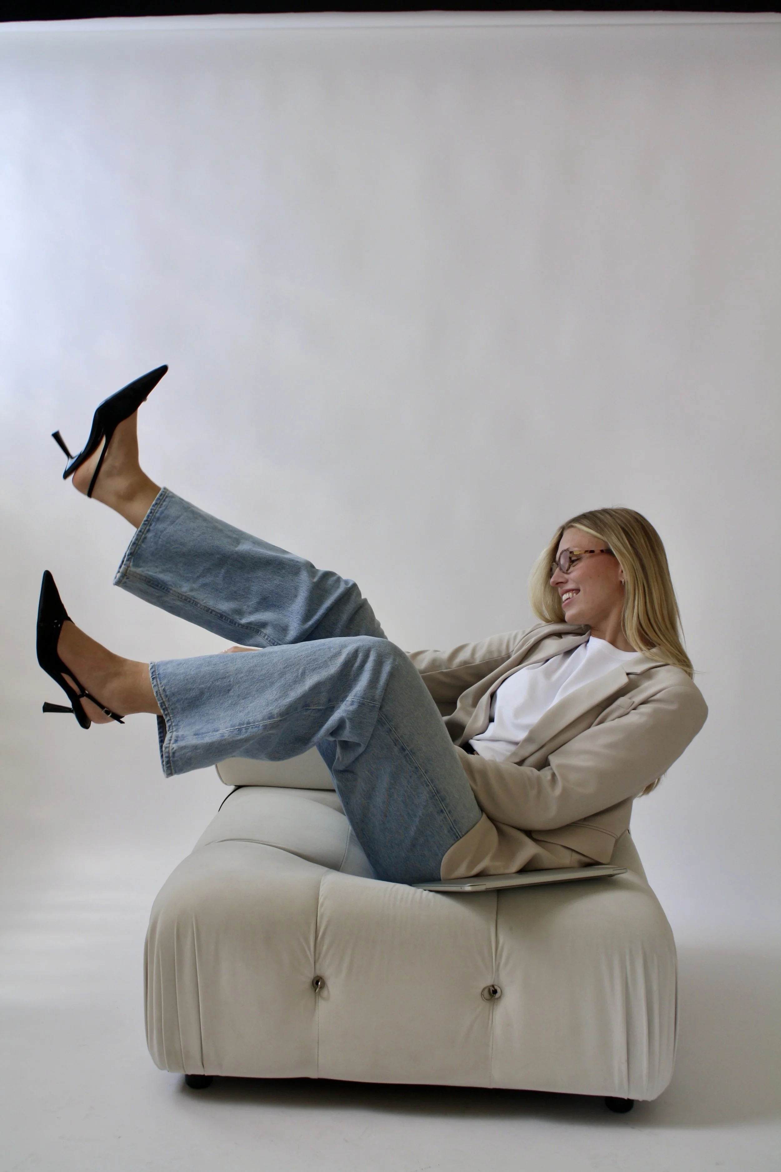 Woman with blonde hair, glasses, and a beige blazer sitting on a white sofa, smiling as she raises her legs, wearing black high heels and light blue jeans, against a plain white background.