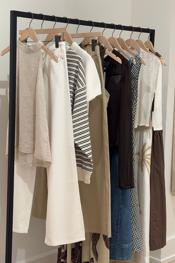 A clothing rack with various beige, white, black, and patterned garments hanging on wooden hangers.
