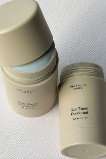 Two sticks of Blue Tansy deodorant with beige packaging, one opened showing the light blue deodorant inside, and the other closed, placed on a white textured surface.