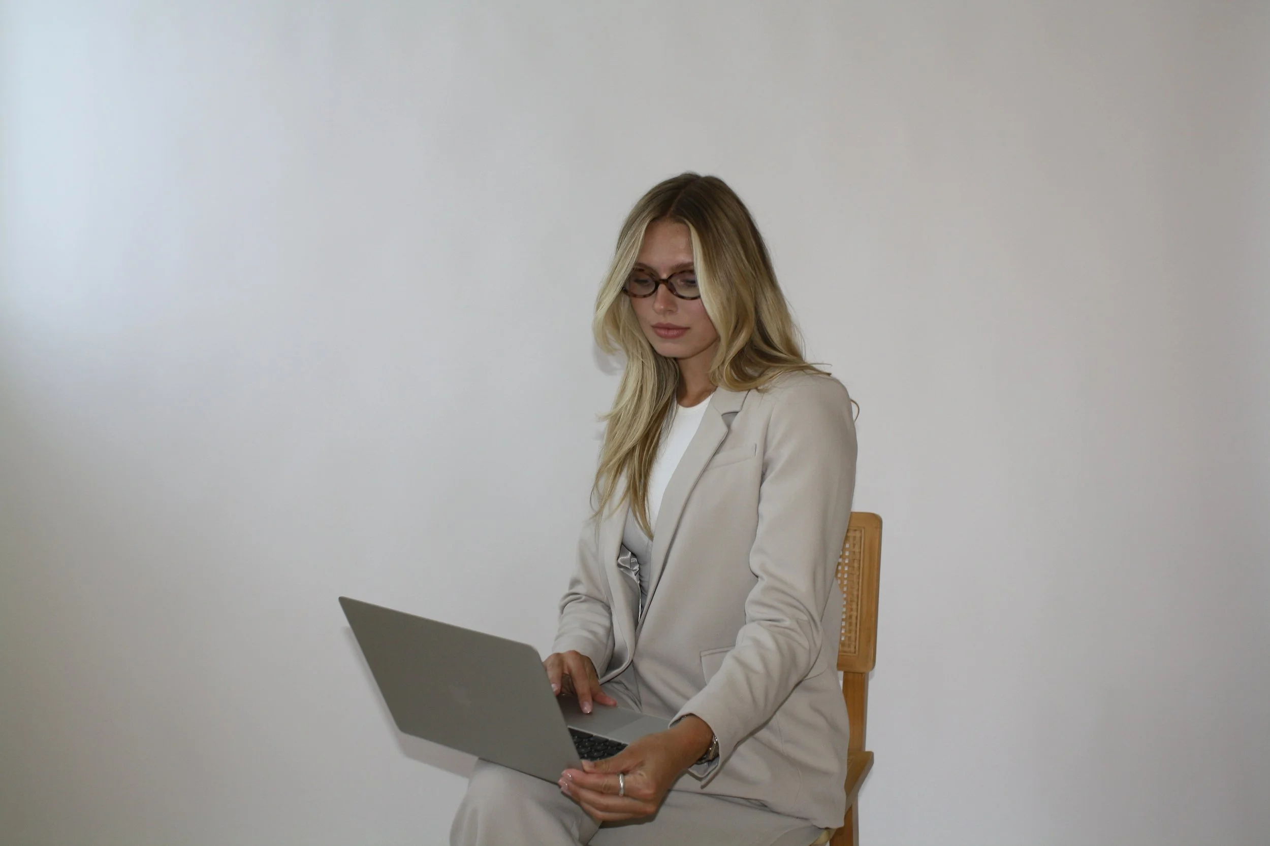 A woman with long blonde hair, wearing glasses and a beige blazer, is sitting on a wooden chair and working on a laptop against a plain white wall background.