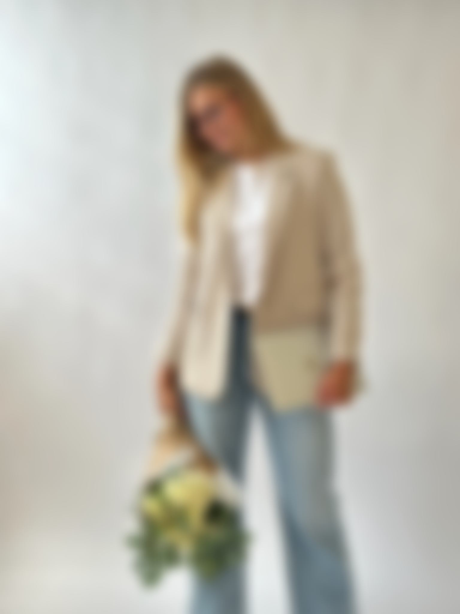 A woman wearing glasses, a white top, tan blazer, and blue jeans, standing and holding a bunch of yellow flowers against a plain light-colored background.