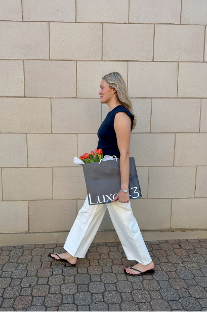 Woman walking on sidewalk carrying a bouquet and shopping bag with tulips in front of a beige brick wall.