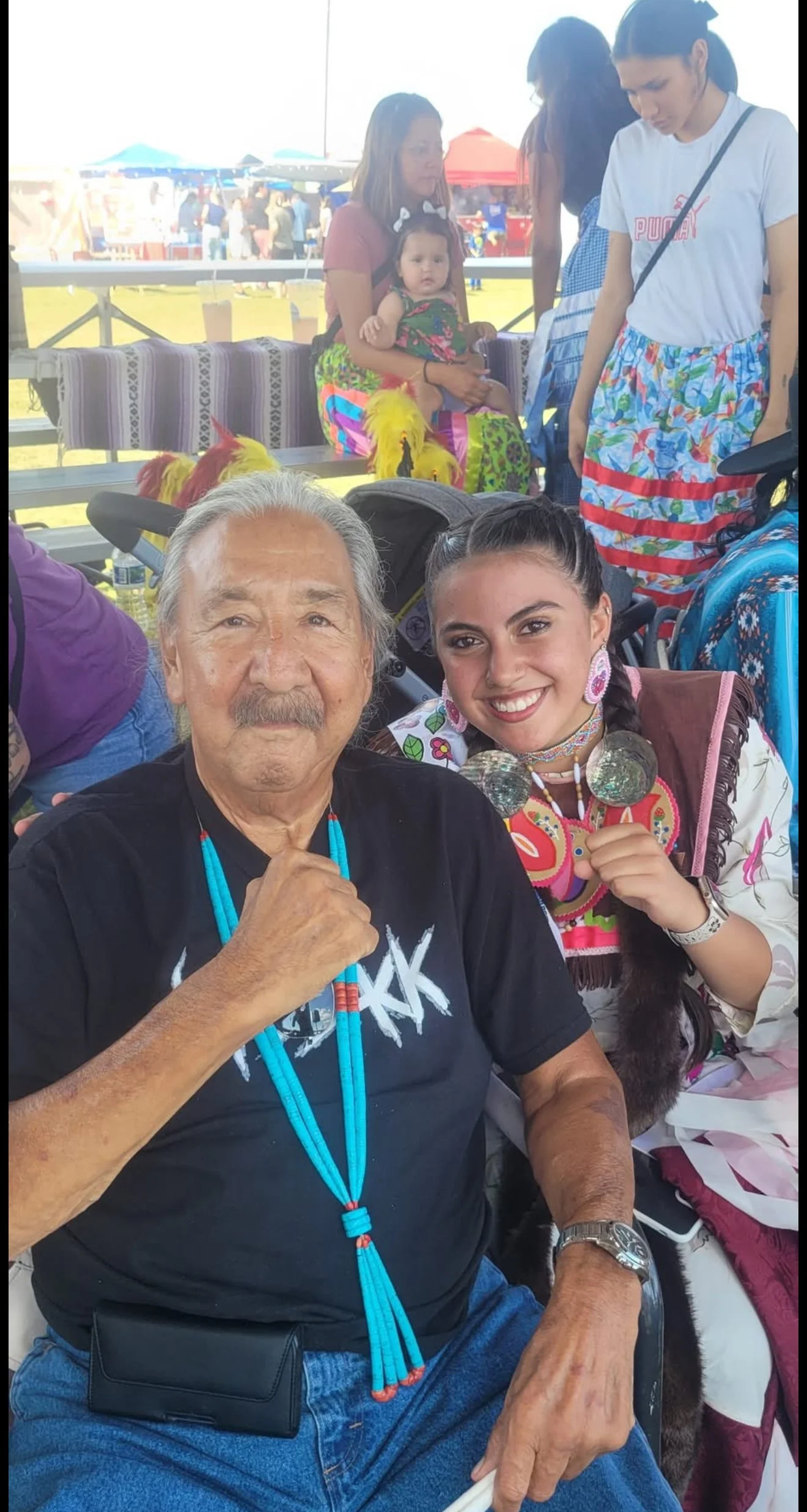 A smiling elderly man and a young woman in colorful traditional attire sitting together at an outdoor event or festival.