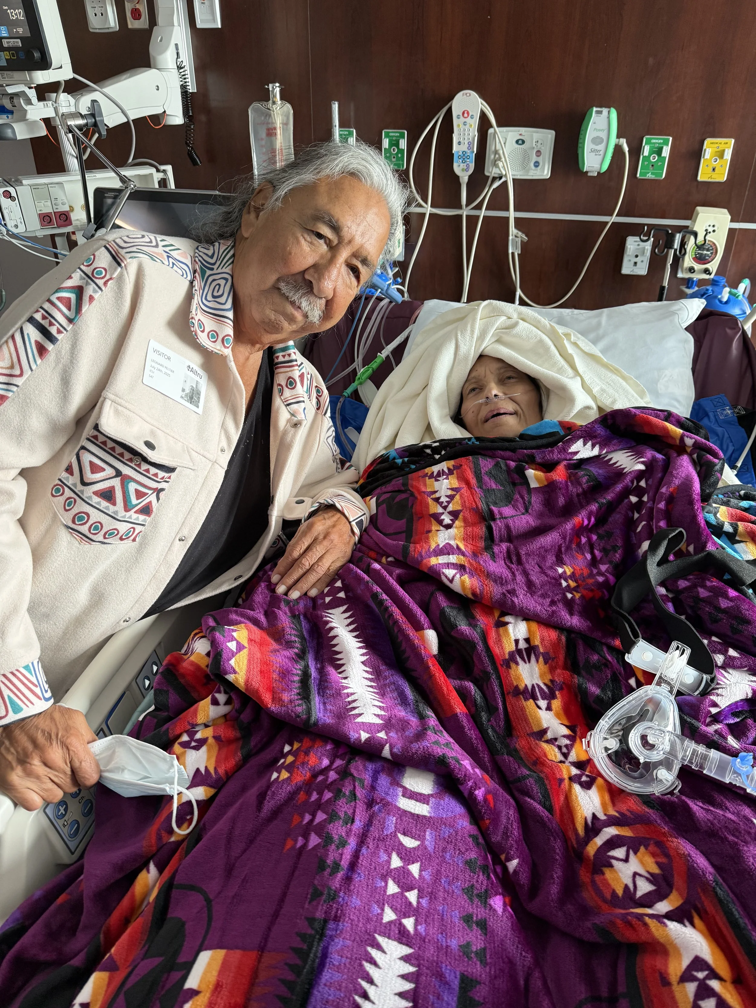An elderly man standing next to a woman lying in a hospital bed, connected to medical equipment. The woman is covered with a colorful patterned blanket and has a nasal oxygen tube.