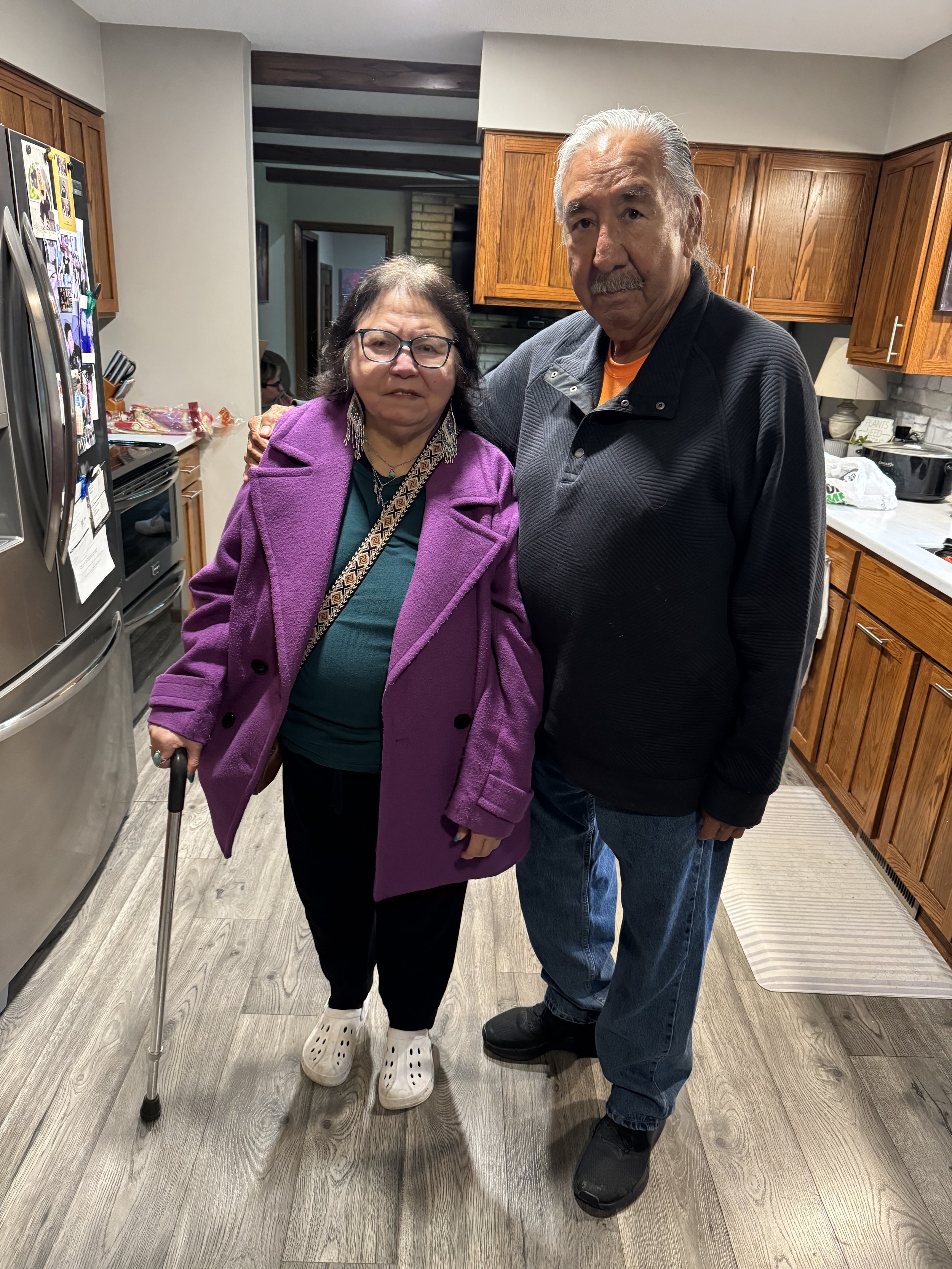 An elderly woman with glasses and a purple coat holding a cane and standing next to an elderly man with white hair and a black jacket, both in a kitchen.