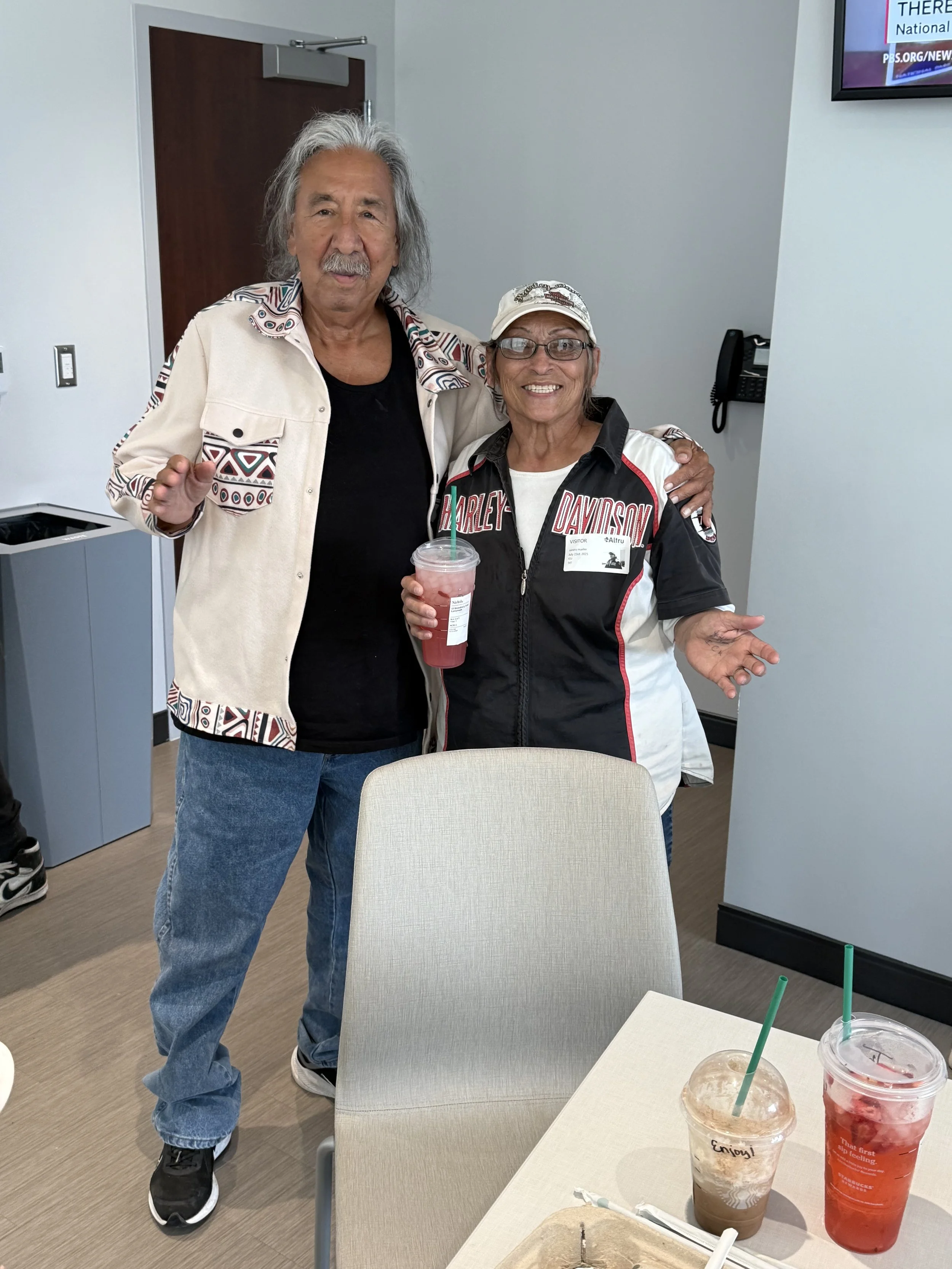 Two people smiling and standing indoors, one wearing a Harley Davidson jacket and the other with long gray hair, holding cold drinks with green straws, in what appears to be a casual setting.