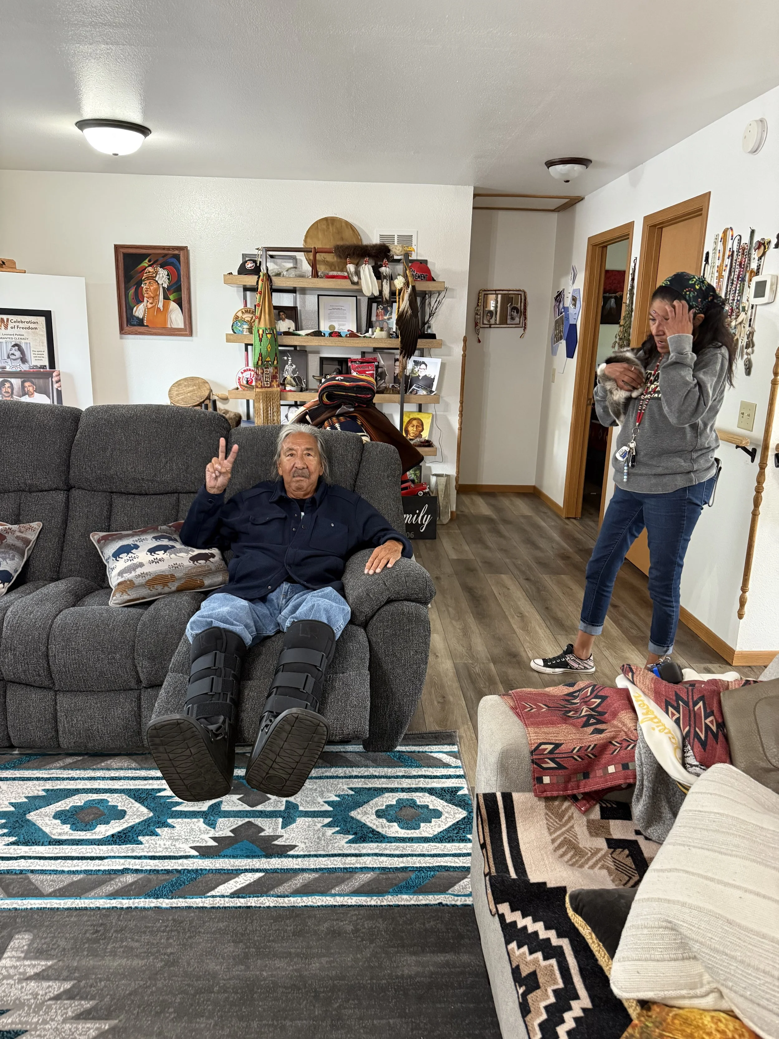 A cozy living room with an elderly man sitting on a gray couch, wearing black leg braces, making a peace sign. A woman stands nearby holding a small dog. The room has decorative pillows, a patterned rug, and a cluttered bookshelf with photos, art, an