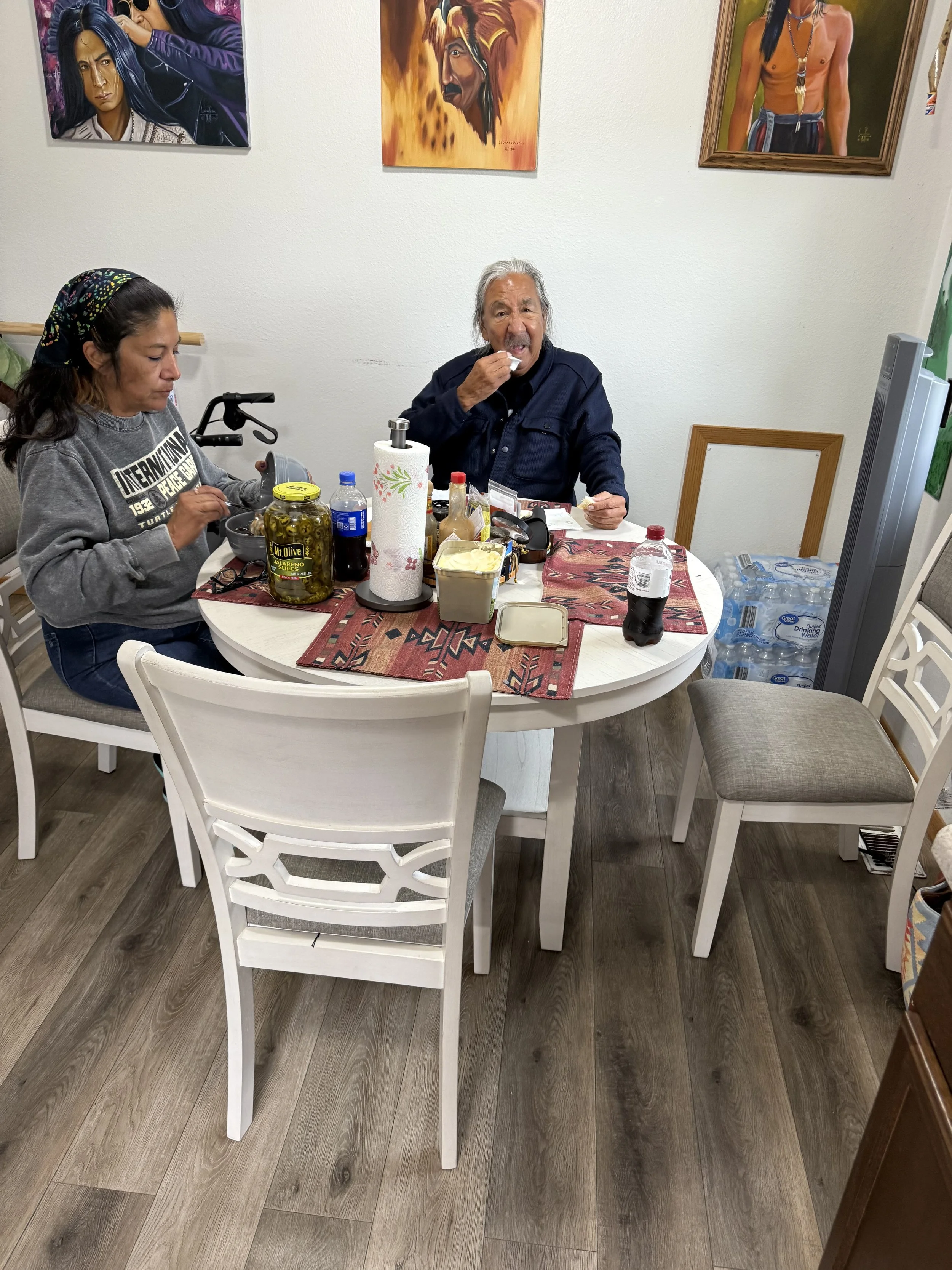 A woman and an elderly man are sitting at a dining table with food and drinks, the woman on the left is wearing a gray sweatshirt and headscarf, and the elderly man on the right is eating a sandwich, there are paintings on the wall behind them, and a