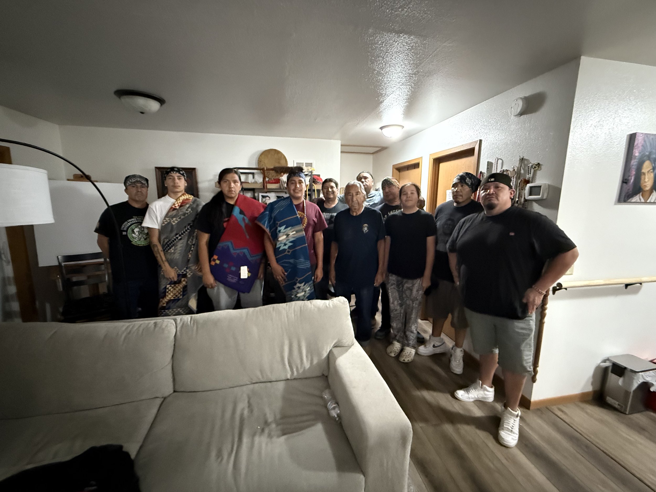 A group of people standing together inside a living room, some dressed in traditional Maori clothing, posing for a photo.
