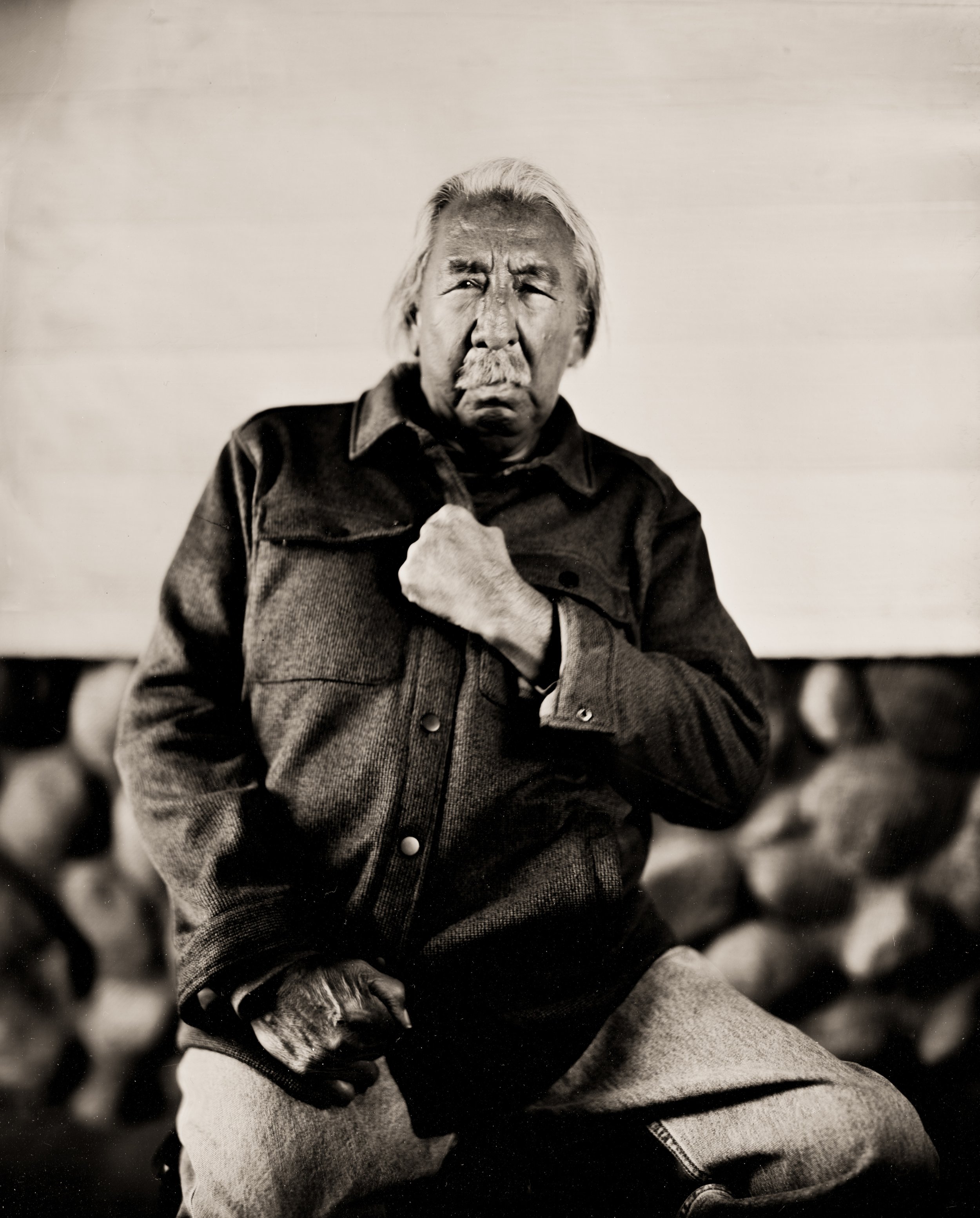 Black and white photo of an elderly Asian man with long white hair and a mustache, making a fist with one hand and holding his jacket with the other, with people blurred in the background.